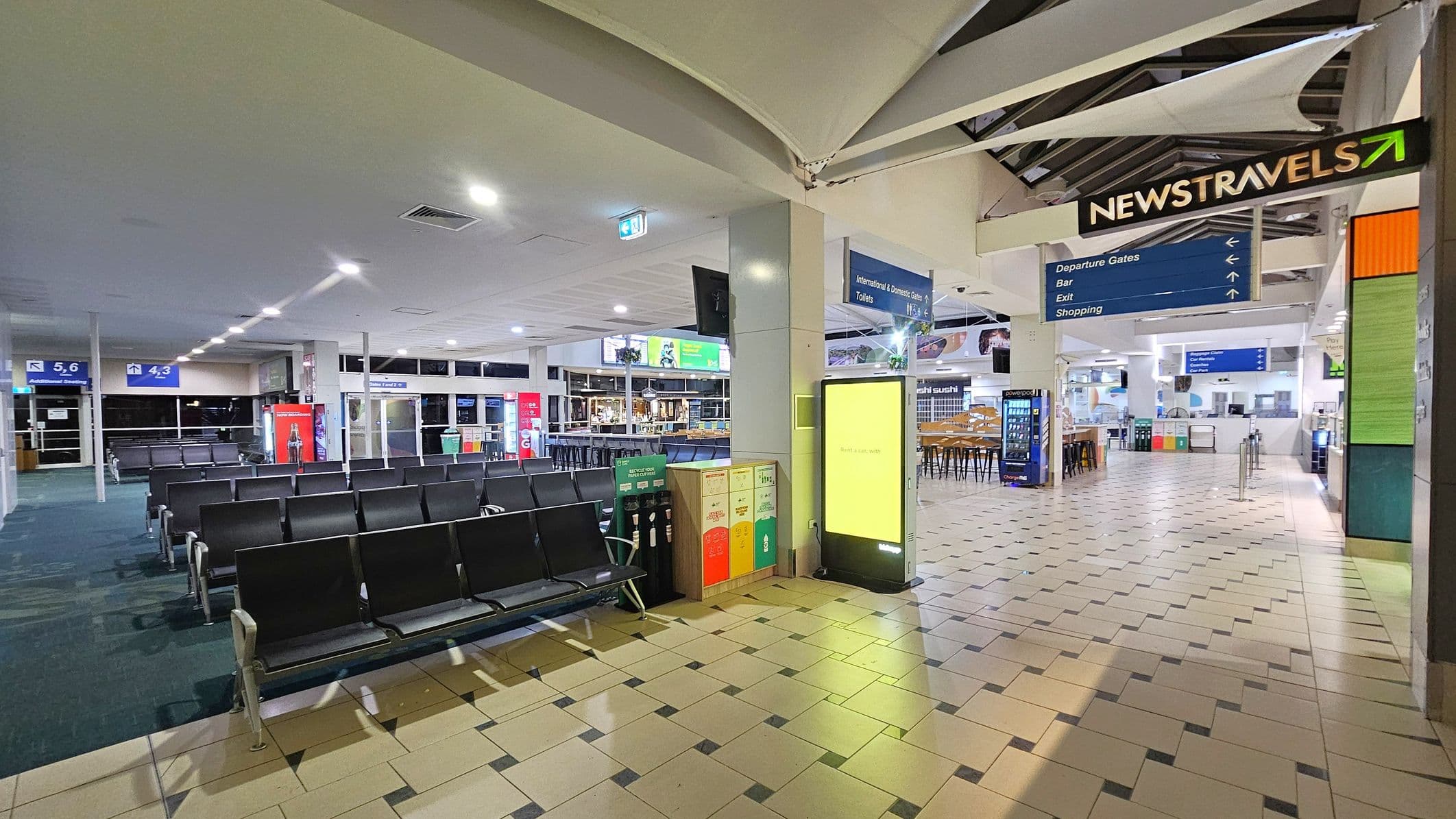 An airport terminal with a tiled floor and a high ceiling. On the left, rows of black seating face towards gates 4, 5, and 6, with vending machines nearby. In the centre, a bright digital display stands next to recycling bins. On the right, a sign for "NEWSTRAVELS" hangs above, with directions for departure gates, a bar, exit, and shopping. Further back, a dining area with tables and chairs is visible, along with more vending machines and signage.