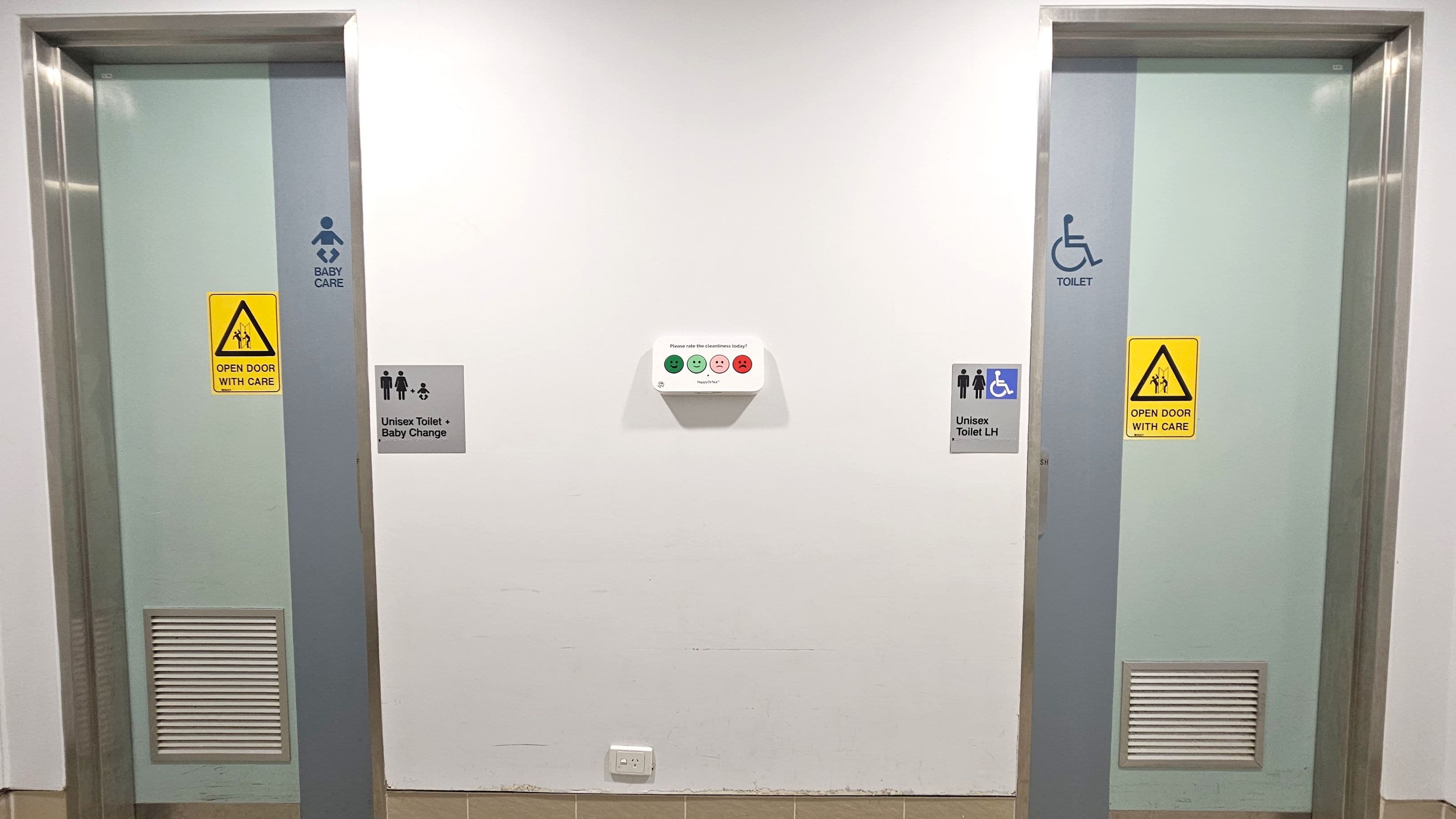 Two doors with metallic frames are set in a white wall. The left door is marked for baby care with a sign and a yellow caution notice. The right door is designated for accessible use, also with a caution sign. Between the doors, a feedback device with smiley faces is mounted on the wall. Below it, there are signs indicating a unisex toilet with baby change on the left and a unisex toilet with left-hand access on the right. The floor is tiled in a light brown colour.