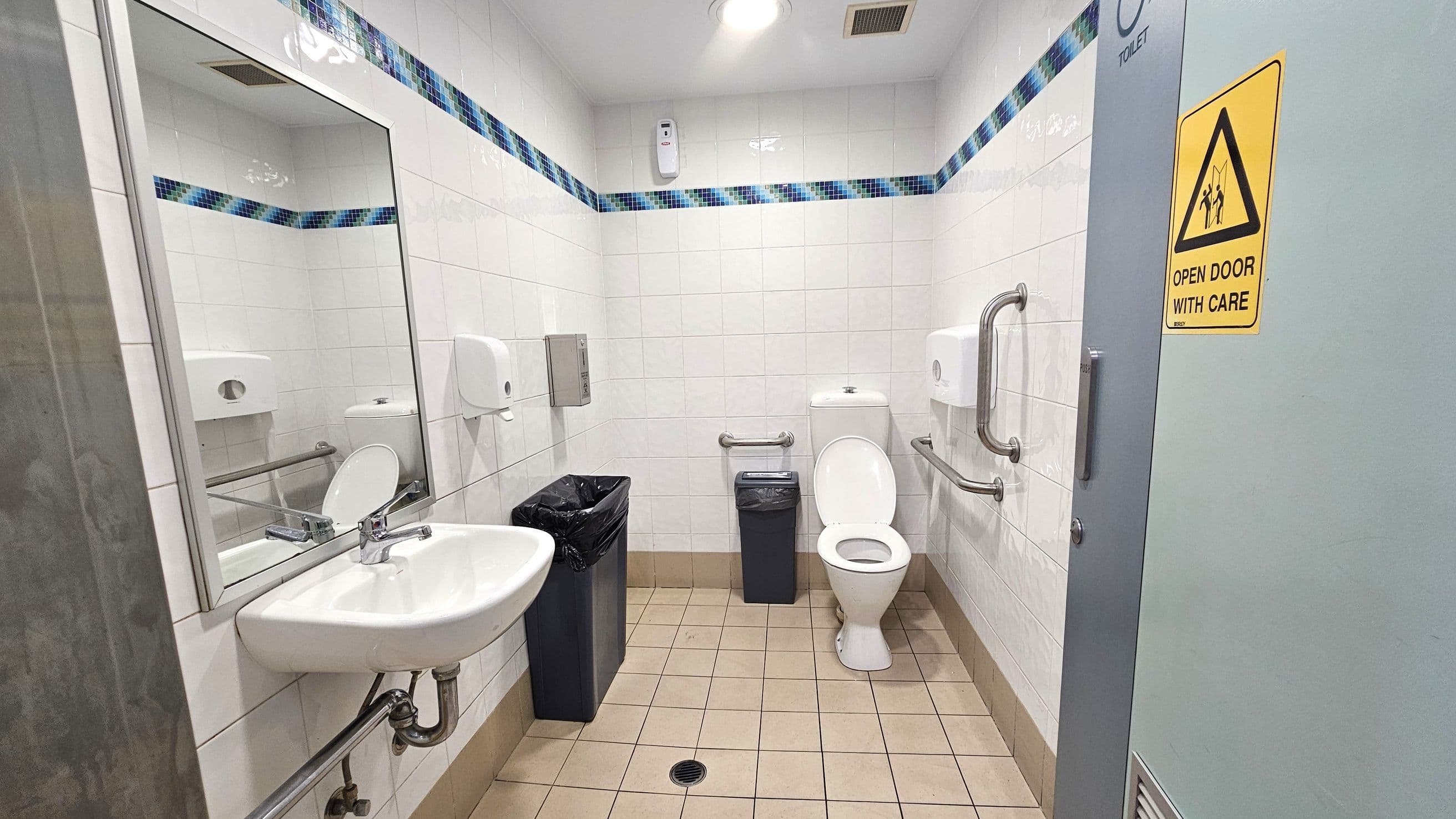 A public restroom with white tiled walls and a blue mosaic border. On the left, a wall-mounted sink with a mirror above it. Below the sink, there are exposed pipes and a metal support bar. In the centre, a white toilet with grab bars on either side and a small bin next to it. On the right, a door with a yellow caution sign reading "Open Door With Care." The floor is covered with beige tiles, and the ceiling has a light fixture and a vent.