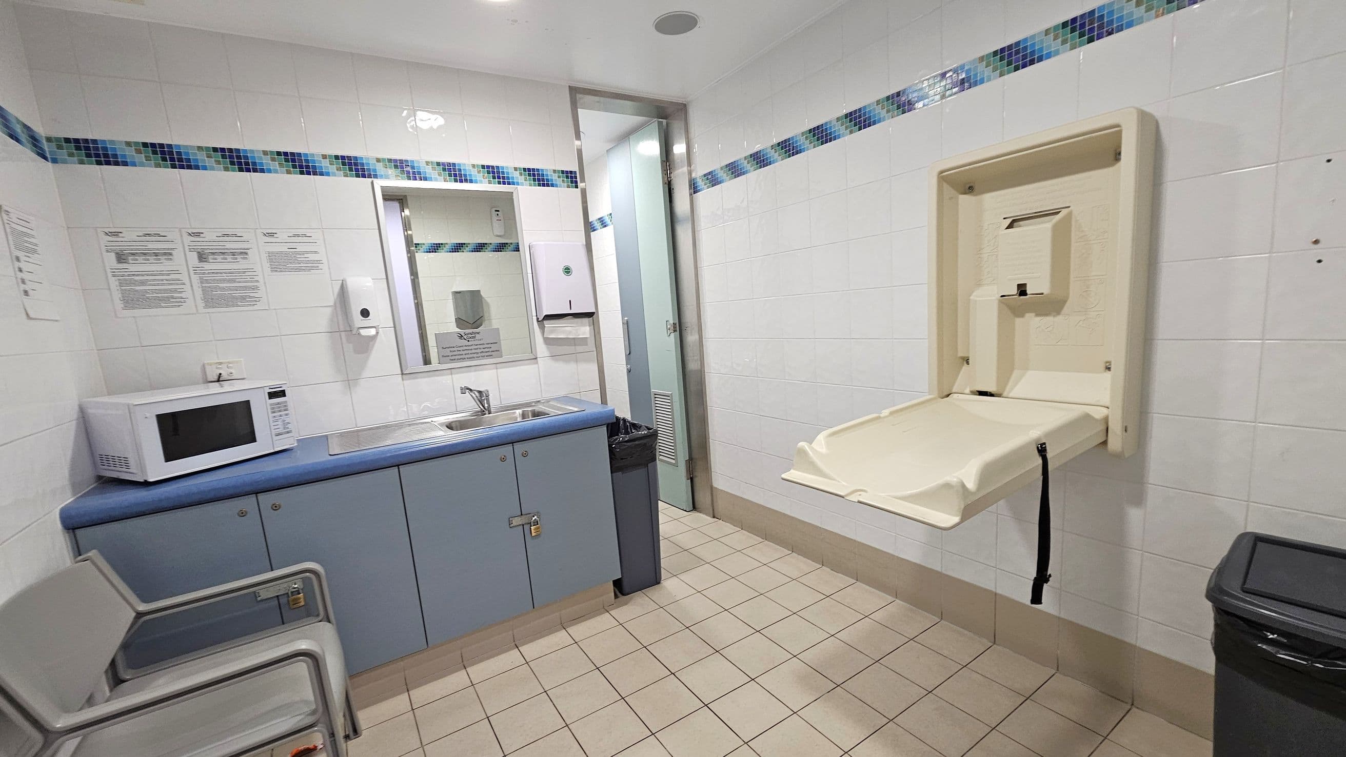 A room with white tiled walls featuring a blue and green mosaic border. On the left, there are two grey chairs, a microwave on a blue countertop, and a sink with a mirror above it. A soap dispenser and paper towel holder are mounted on the wall. In the centre, a door leads to another area. On the right, a baby changing station is mounted on the wall. The floor is covered with beige tiles, and a black bin is positioned near the changing station. The ceiling is white with recessed lighting.