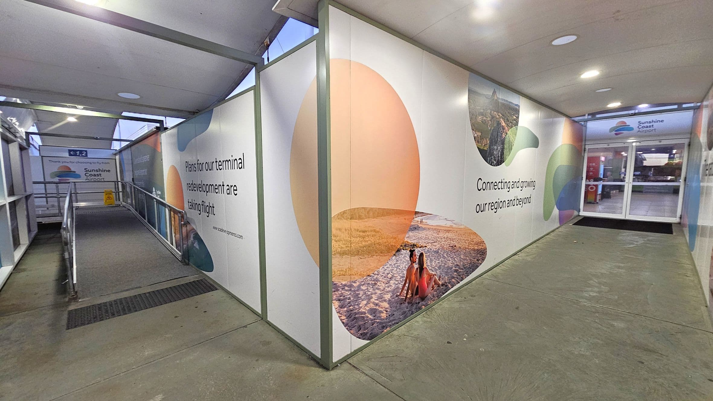 A corridor with a ramp on the left leading to a sign that reads "Sunshine Coast Airport." The walls are adorned with promotional graphics and text about terminal redevelopment. On the right, there is a glass door entrance. The floor is concrete, and the ceiling is covered with panels featuring recessed lighting.