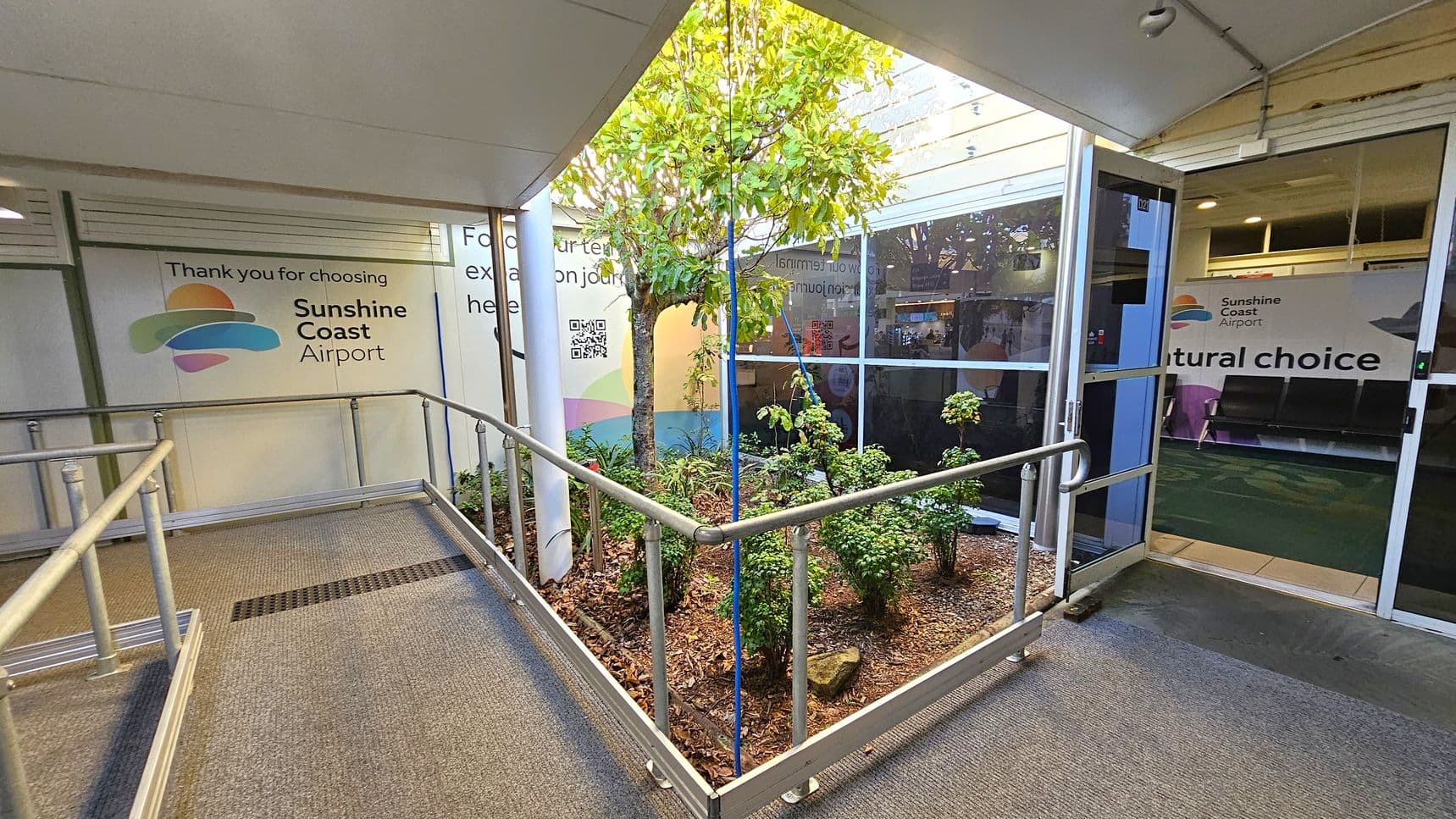 A corridor with a grey carpeted floor and metal railings on both sides. On the left, a wall displays the Sunshine Coast Airport logo and a message. In the centre, a small garden area with a tree and shrubs is enclosed by railings. On the right, a glass door leads to an interior space with seating and another Sunshine Coast Airport sign. The ceiling is plain and white.