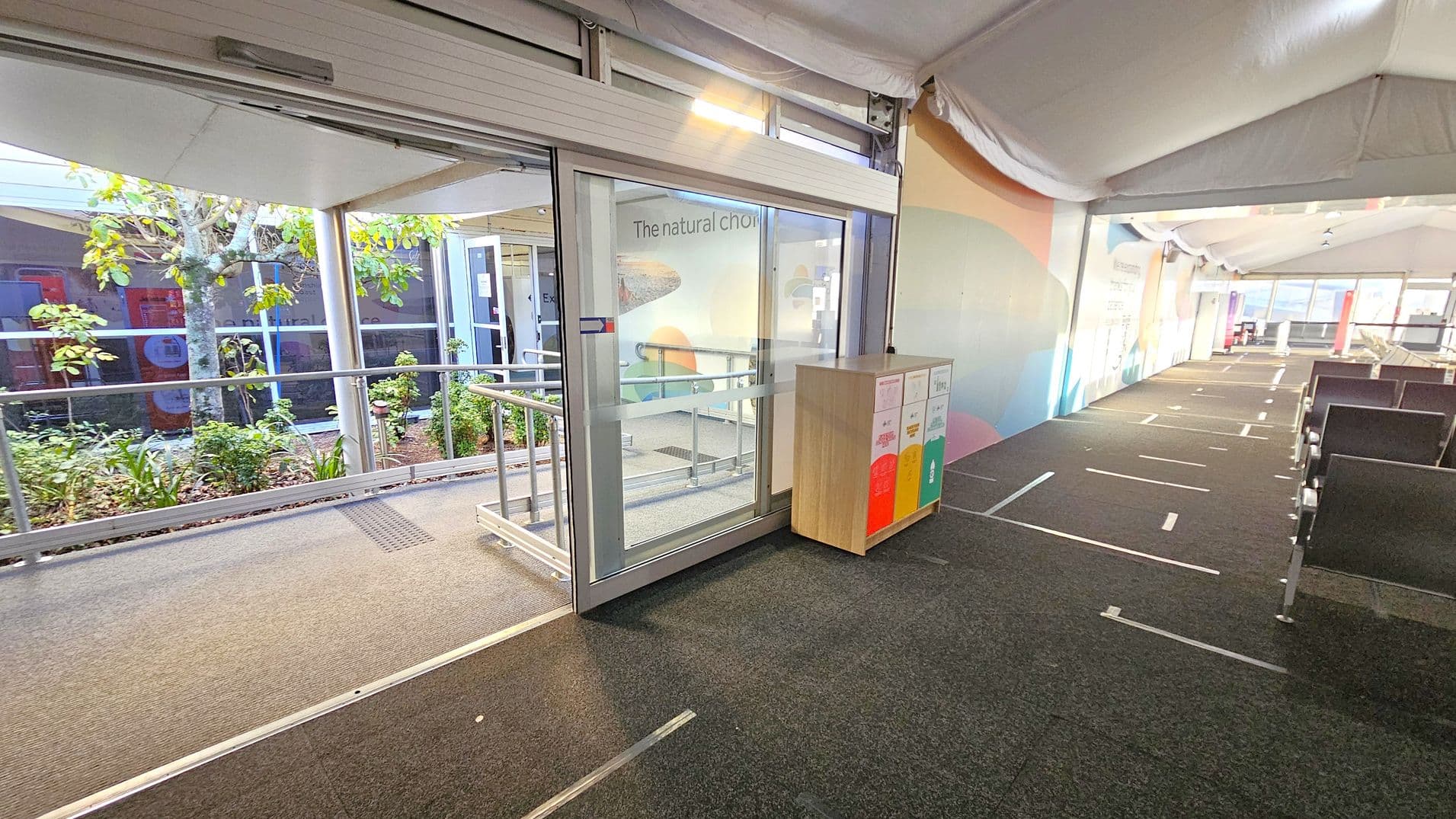 A corridor with a grey carpeted floor and a white tent-like ceiling. On the left, there is a glass door leading outside, with a view of plants and a tree. A wooden recycling bin with colourful labels is positioned near the door. On the right, rows of black chairs are aligned along the wall, which features a colourful mural. The corridor extends into the distance, with more seating visible further along.