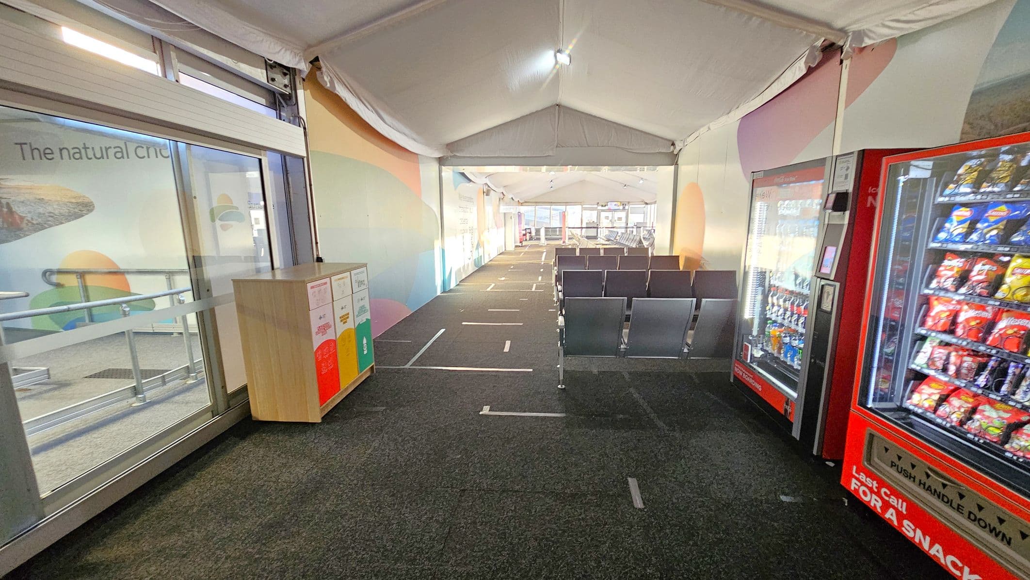 A corridor with a grey carpeted floor and a white tent-like ceiling. On the left, there's a glass door with a colourful recycling bin nearby. The wall has a mural with soft colours. On the right, two red vending machines are filled with snacks and drinks. Further down, rows of grey metal chairs are arranged in a waiting area. The space is well-lit with natural light coming from windows at the far end.