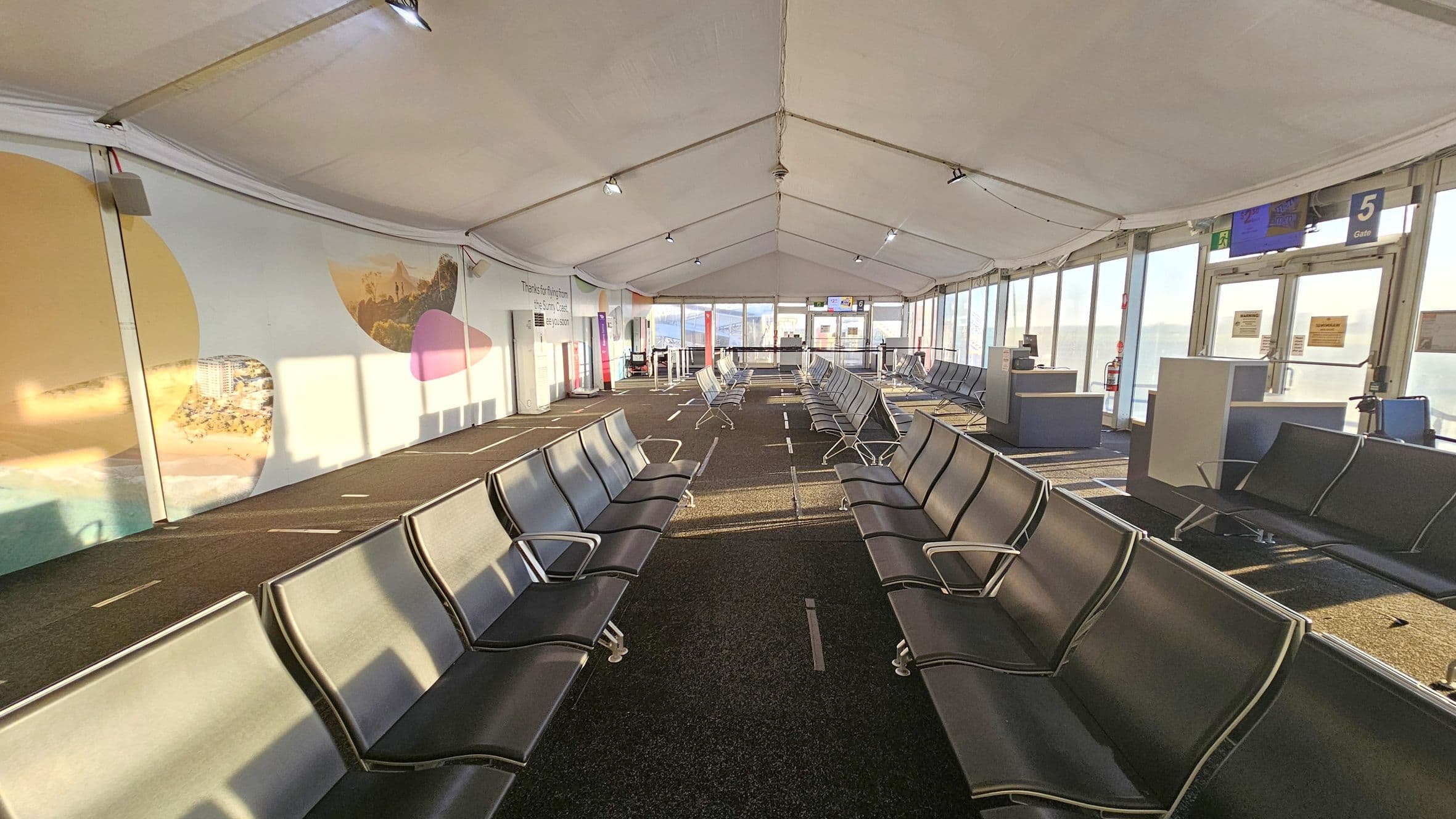 A spacious airport waiting area with rows of black seating on both sides. The left wall features large, colourful murals with abstract shapes and images of landscapes. The right side has large windows allowing natural light to flood in, with signs for Gate 5 visible. The floor is carpeted in dark tones, and the ceiling is tent-like with exposed beams and lighting fixtures. In the background, there are more seats and a boarding area with signs and barriers.