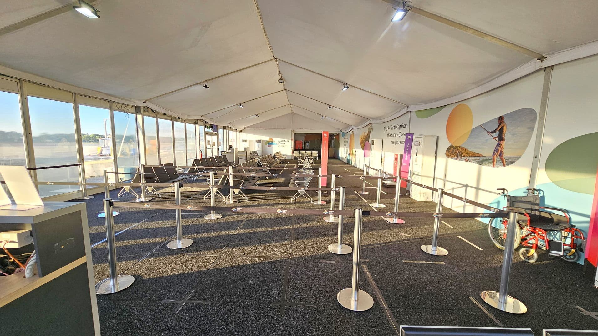 Inside an airport terminal with a white tent-like ceiling. On the left, there are large windows allowing natural light to illuminate the space. In the centre, rows of grey seating are arranged, with metal stanchions forming queue lines. The right wall features colourful murals and a sign thanking travellers. A red wheelchair is positioned near the wall. The floor is carpeted in dark grey, and overhead lights are evenly spaced along the ceiling.