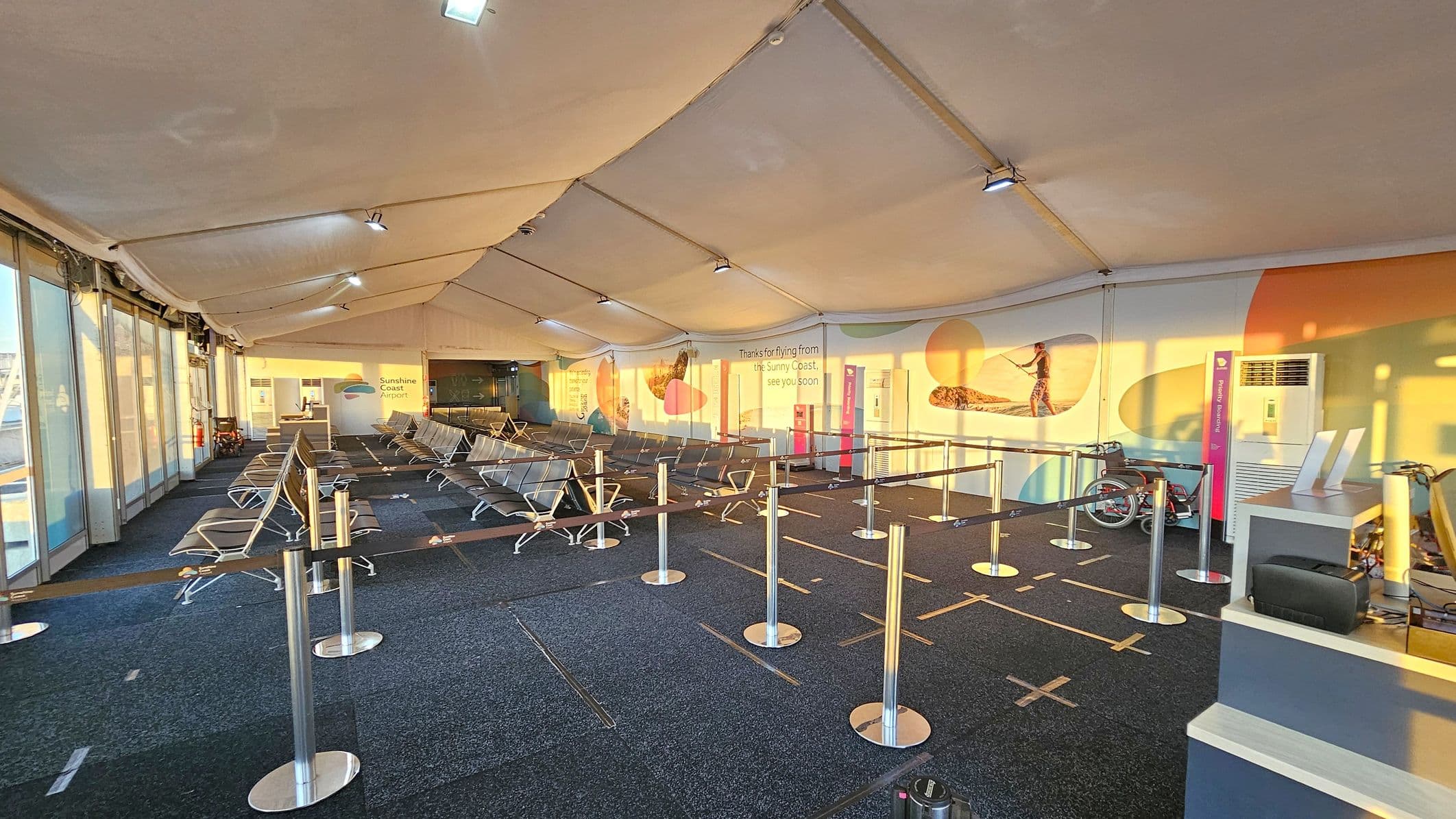 Inside an airport terminal with a high, tent-like ceiling. On the left, there are large glass doors and rows of metal seating. The right side features a counter with electronic devices and a wheelchair nearby. The back wall displays colourful graphics and text reading "Thanks for flying from the Sunny Coast, see you soon." The floor is carpeted with a dark pattern, and there are stanchions with belts forming a queue area. Bright lighting creates a warm atmosphere.