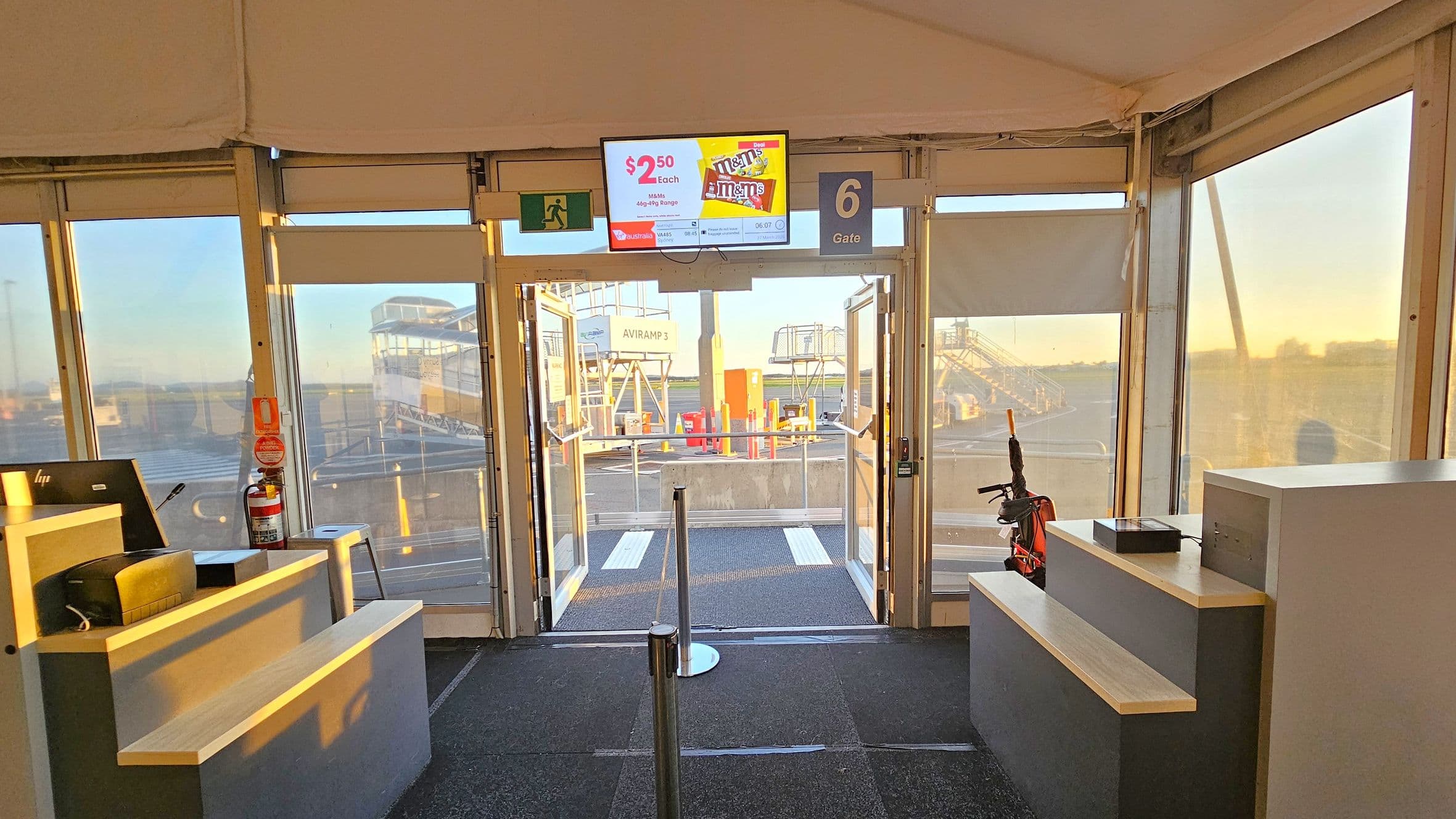 Inside an airport gate area with a carpeted floor. In the centre, glass doors lead to the tarmac, where boarding ramps are visible. Above the doors, a digital screen displays an advertisement. To the left, a counter with a computer and a fire extinguisher. On the right, another counter with a small device and an umbrella. The ceiling is tent-like, and the area is well-lit with natural light streaming through the windows.