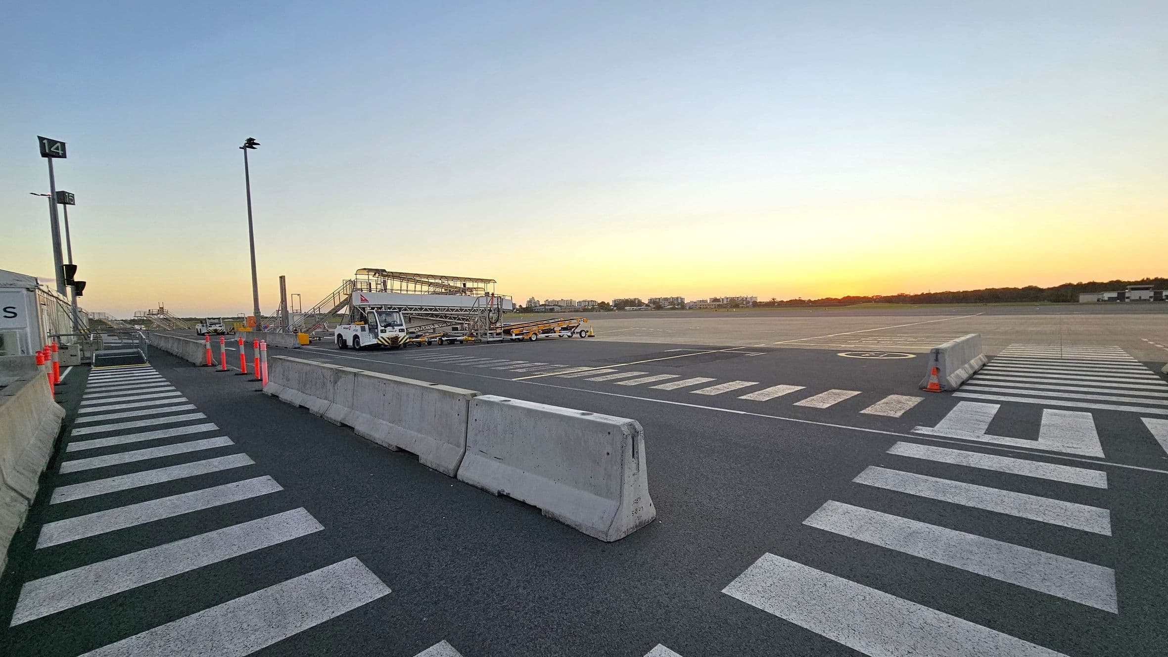 A tarmac area with a wide view of an airport apron. On the left, there are numbered signs and a pedestrian walkway marked with white stripes. Concrete barriers separate the walkway from the main tarmac. In the centre, there are mobile staircases and ground service vehicles. The right side shows an open expanse of tarmac with a few distant buildings and trees on the horizon. The sky is clear with a warm glow near the horizon.