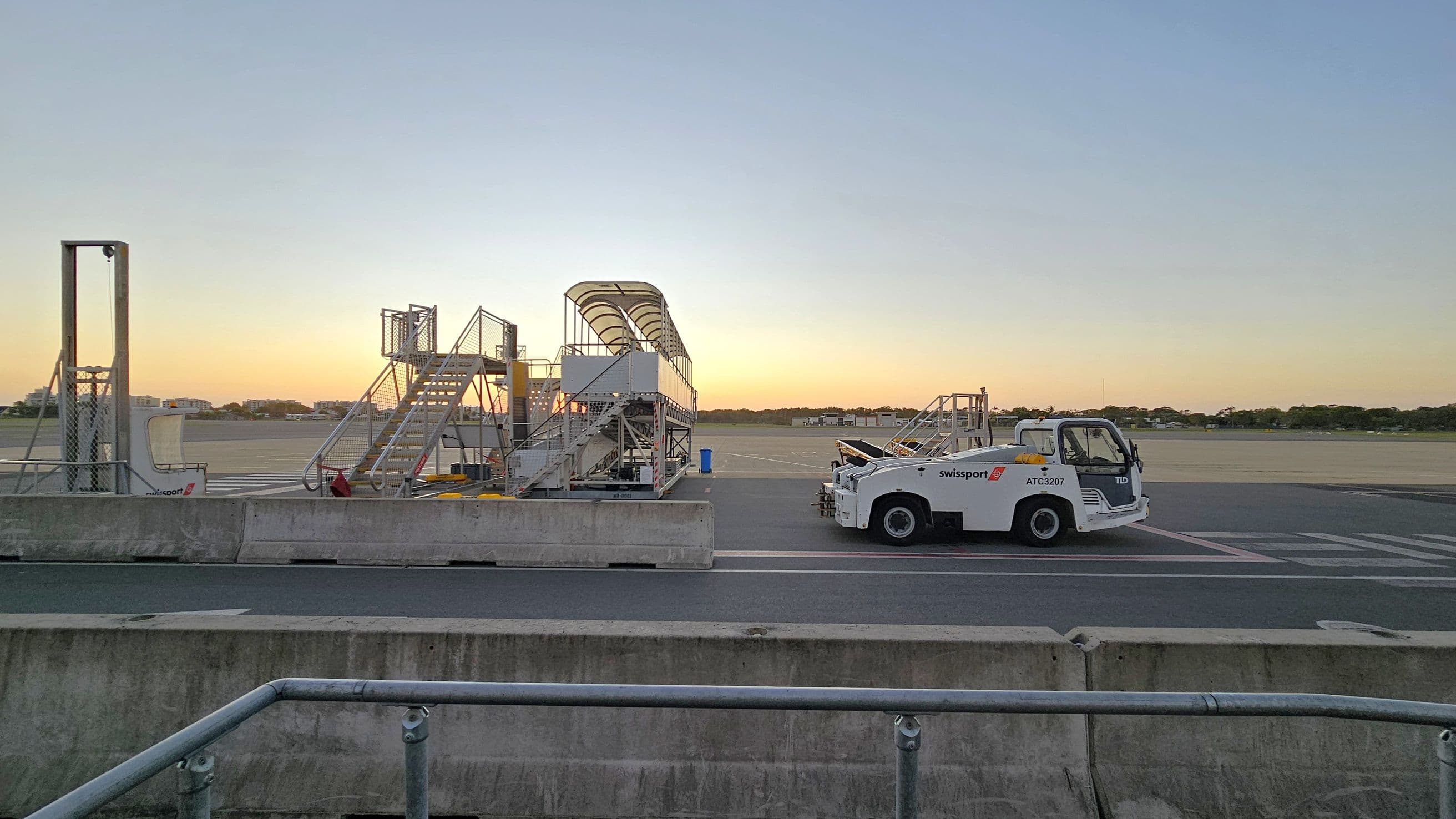 On the left, there is a set of mobile stairs with a canopy, positioned on an airport tarmac. To the right, a white airport vehicle marked "Swissport" is parked beside another set of mobile stairs. In the background, the tarmac extends towards a line of trees and distant buildings. The foreground features a concrete barrier with a metal railing. The sky is clear with a gradient from light to darker tones.