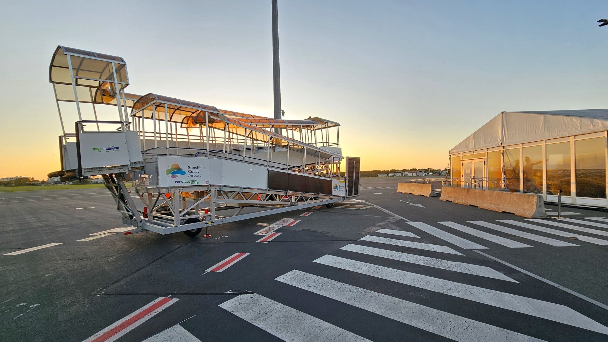 A mobile passenger boarding ramp is positioned on an airport tarmac. The ramp is labelled with "Sunshine Coast Airport" and "100% Electric". It has a covered walkway with a metal frame. To the right, there is a large white tent-like structure with glass windows, and concrete barriers in front. The ground features a marked pedestrian crossing with white stripes. The background shows an open area with a clear sky and a hint of sunset.
