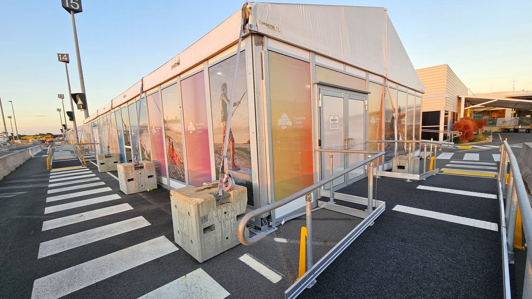 A temporary structure with transparent panels and colourful images on the sides, labelled "Sunshine Coast Airport". The entrance has a metal ramp with railings. Concrete blocks with straps secure the structure. The ground is marked with white pedestrian crossing lines. In the background, there are numbered poles and a building with a modern design. The area is well-lit, and the sky is clear.