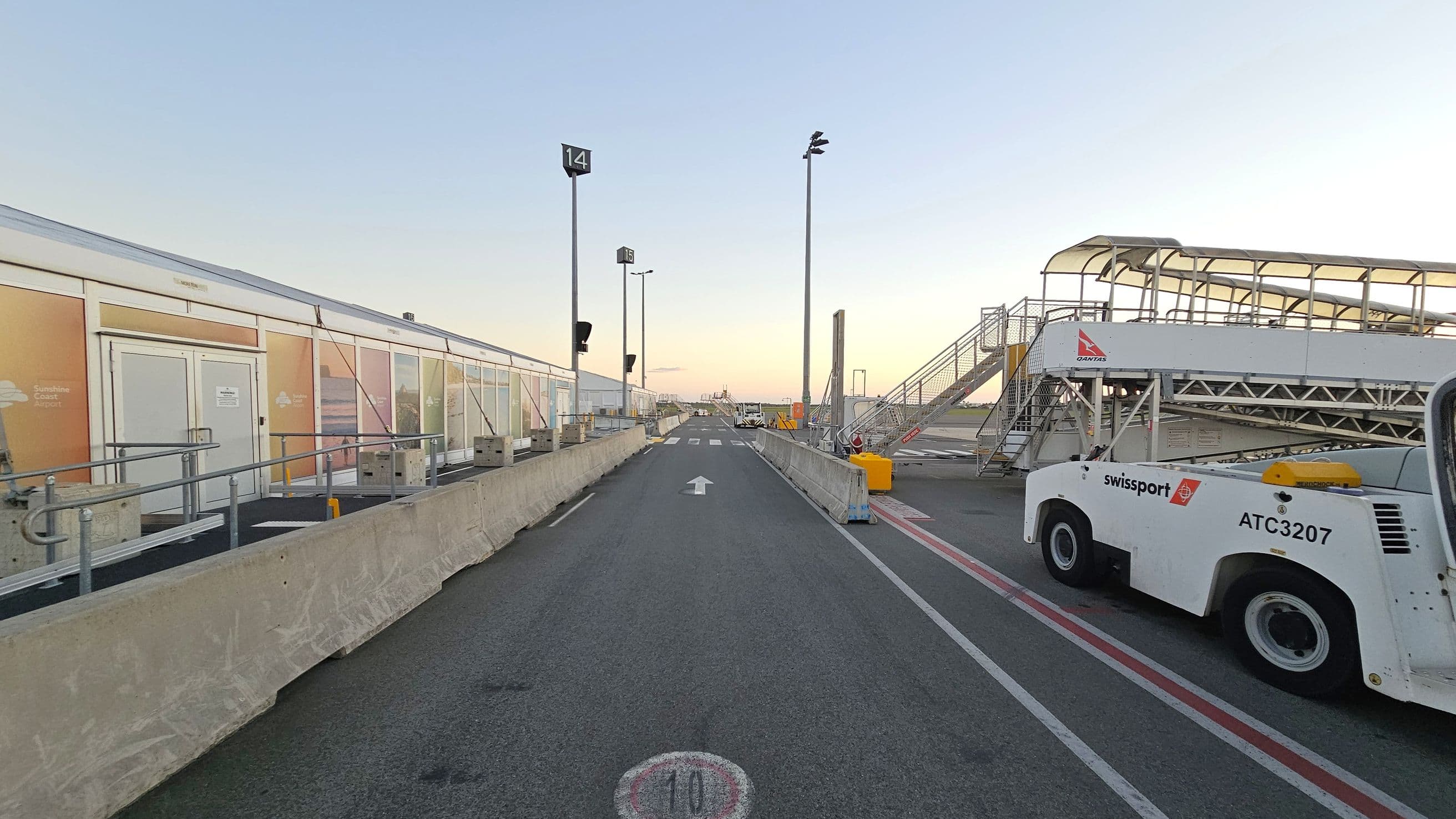 A tarmac area with a road leading into the distance, marked with an arrow and a speed limit of 10. On the left, there are temporary structures with railings and signage. On the right, a Swissport vehicle is parked near a mobile staircase with a Qantas logo. The area is bordered by concrete barriers, and there are tall light poles along the road. The sky is clear, and the scene is well-lit.