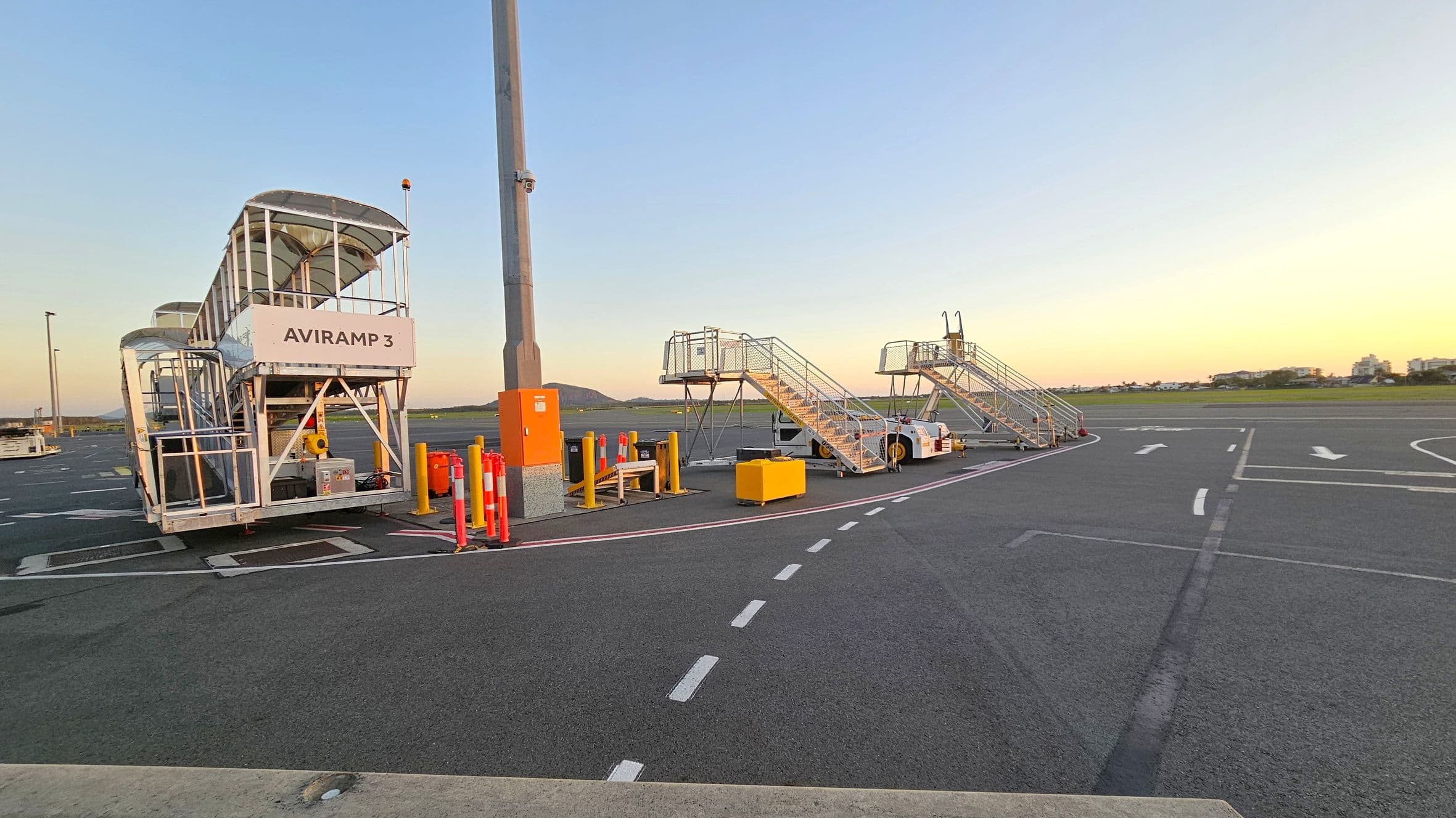 On the left, there is a covered mobile ramp labelled "AVIRAMP 3" with a metal frame and canopy. In the centre, a tall light pole stands next to orange and yellow safety barriers. On the right, two open metal staircases are positioned on the tarmac, each attached to a small vehicle. The ground is marked with white dashed lines and arrows. In the background, there is a grassy area and distant buildings under a clear sky.