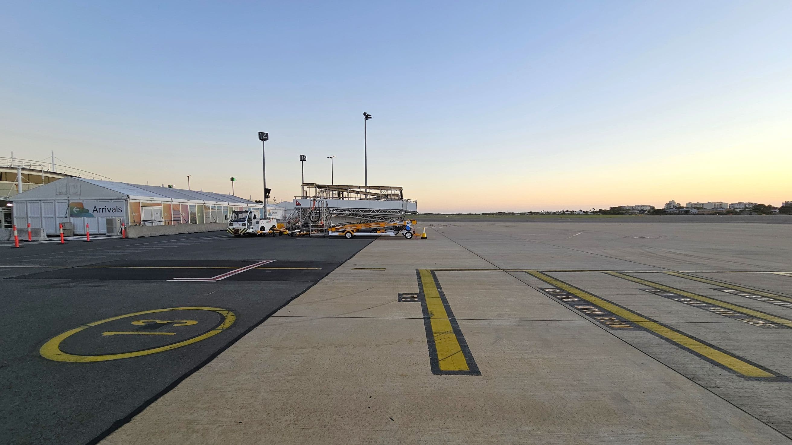 On the left, there is a long, white airport terminal building with a sign indicating "Arrivals". Several orange traffic cones are lined up along the building. In the centre, there is a vehicle with a mobile staircase attached, parked on the tarmac. The ground is marked with various lines and numbers, including a large yellow circle with a number inside. On the right, the expansive tarmac stretches towards the horizon, with a few buildings visible in the distance.