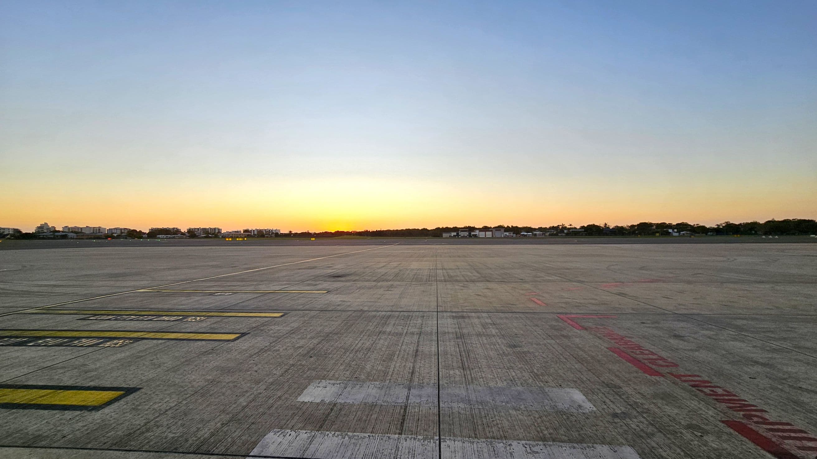 A wide, empty airport runway stretches across the foreground with various markings in yellow and red. In the background, a line of trees and low buildings is silhouetted against a sunset. The sky transitions from a warm orange near the horizon to a pale blue higher up. The surface of the runway is textured with visible lines and grooves.