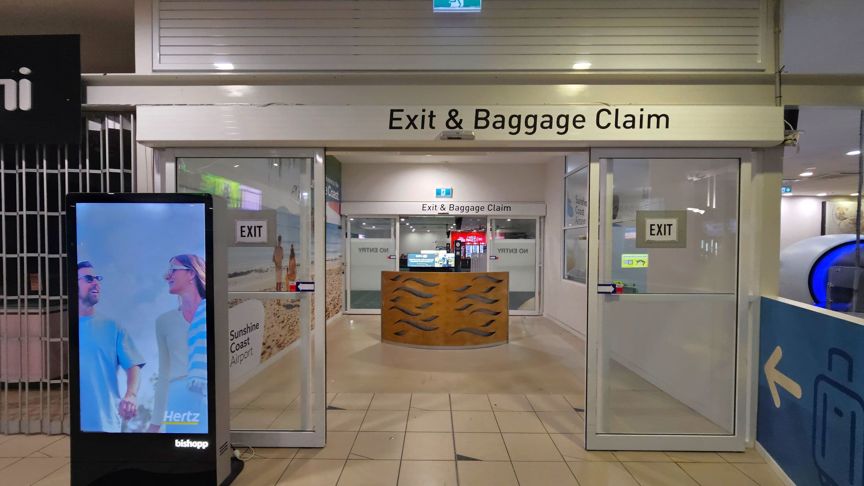 A corridor leading to the "Exit & Baggage Claim" area. On the left, there's a digital advertisement display showing a man and woman smiling. The entrance has glass doors with "EXIT" signs. Inside, a wooden counter with wave patterns is visible. The floor is tiled, and the ceiling has recessed lighting. To the right, there's a blue sign with a luggage icon and an arrow pointing towards the exit.