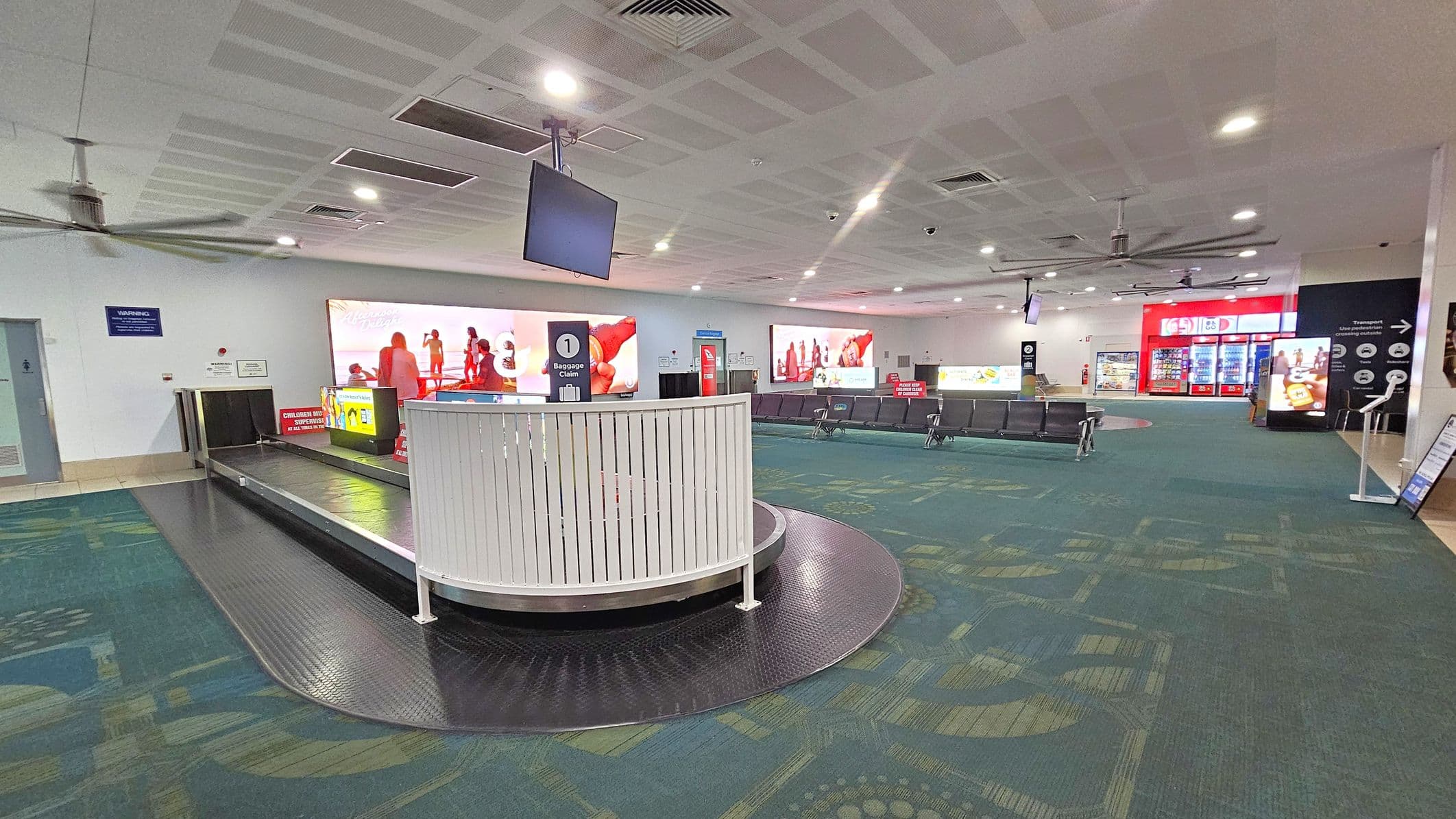 A spacious airport baggage claim area with a conveyor belt in the centre, surrounded by a white railing. To the left, there's a wall with a warning sign and a ceiling fan. On the right, there are rows of seating and a bright red vending machine area. The floor is carpeted in a patterned green and blue design. The ceiling is white with recessed lighting and multiple ceiling fans. Large advertisement screens are visible in the background.