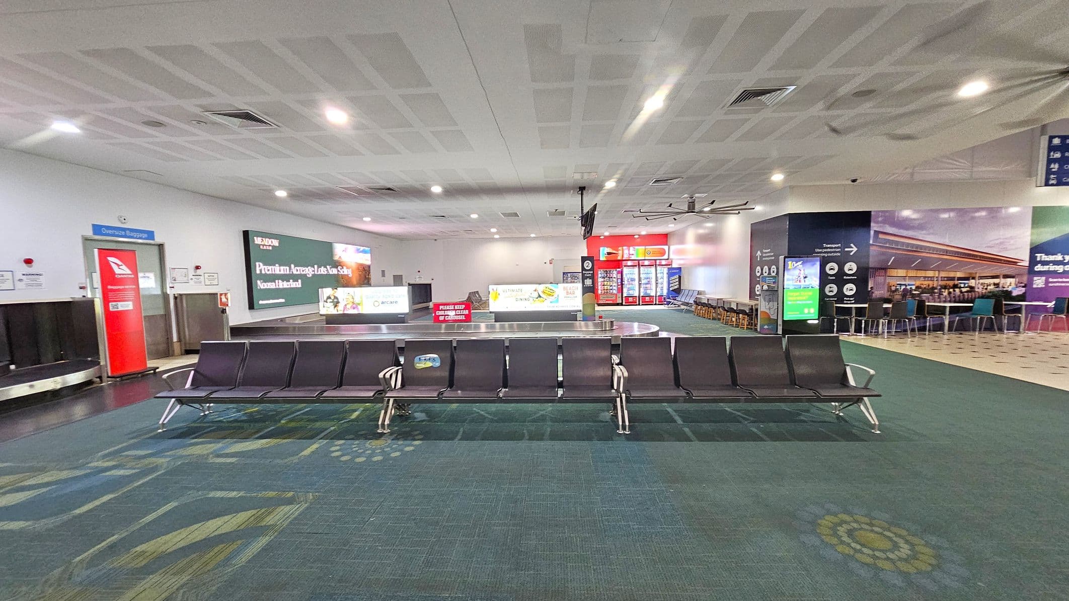 Rows of black airport seating are in the foreground on a patterned green carpet. Behind them is a baggage carousel with a large advertisement on the wall above it. To the left, there's a red door marked "Oversize Baggage". On the right, vending machines and a seating area with tables and chairs are visible. The ceiling is white with recessed lighting and air vents.