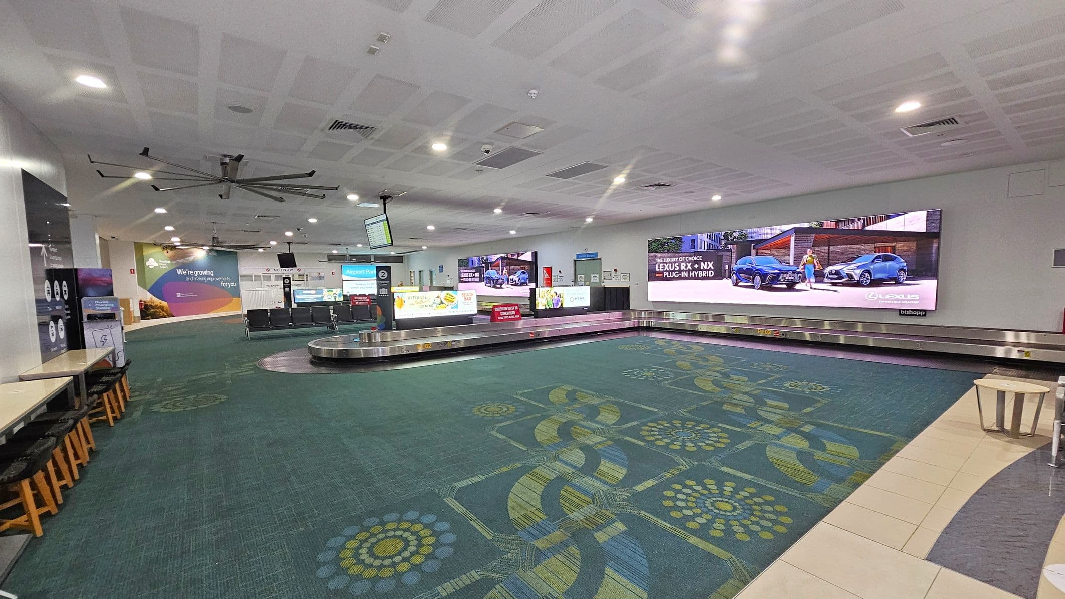 A spacious airport baggage claim area with a patterned green carpet covering the floor. On the right, there is a long baggage carousel with a large digital advertisement above it. In the centre, there are several rows of seating and a digital information screen hanging from the ceiling. On the left, there are tables with stools and various signs. The ceiling is white with recessed lighting and a large ceiling fan.