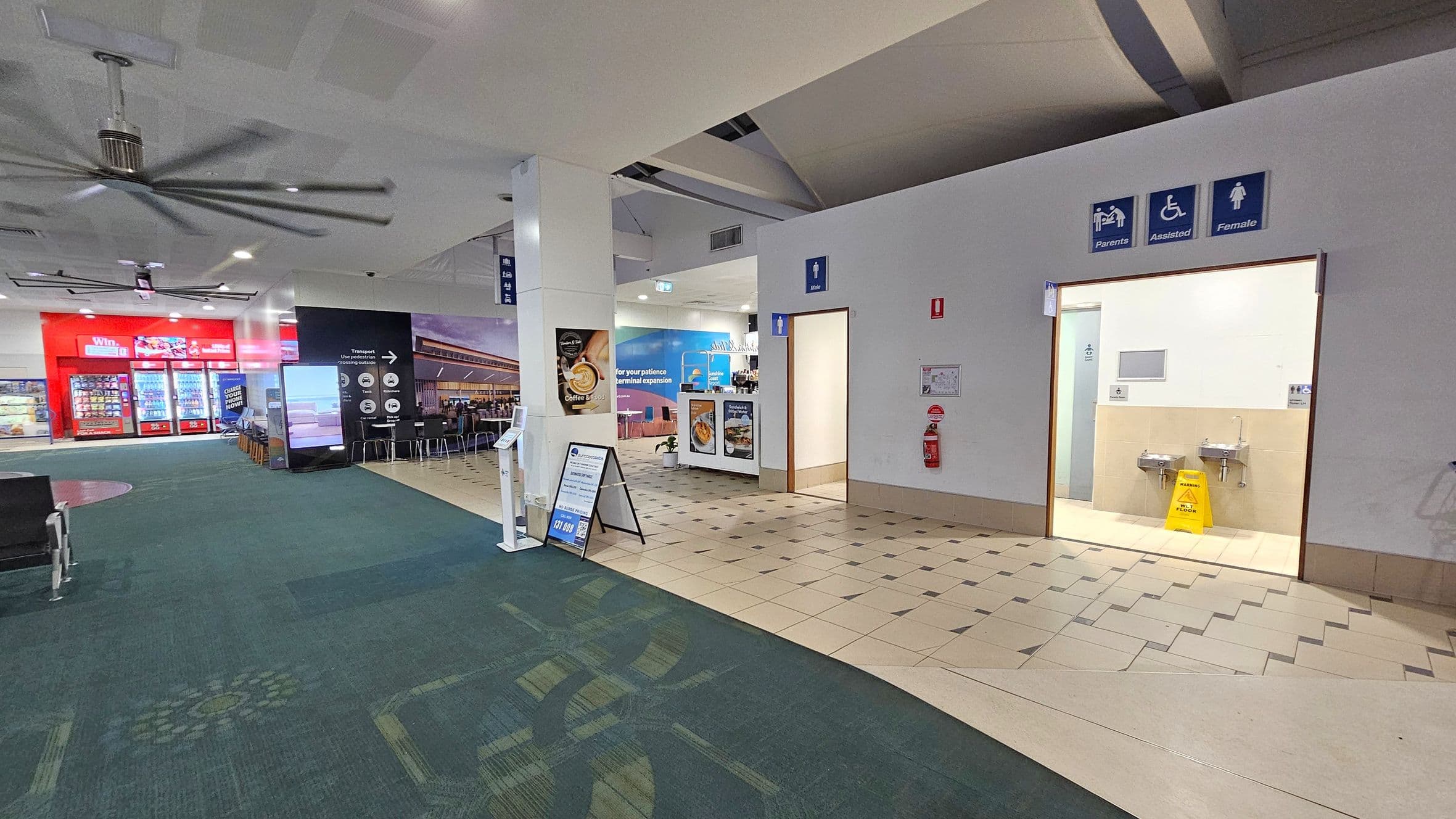 On the left, there are vending machines with bright red signage and a seating area with tables and chairs. The floor is carpeted in green with a pattern. In the centre, a large white pillar displays a sign for coffee. On the right, there are entrances to restrooms with signs for parents, assisted, and female facilities. The floor transitions to beige tiles near the restrooms. A large ceiling fan is visible above.