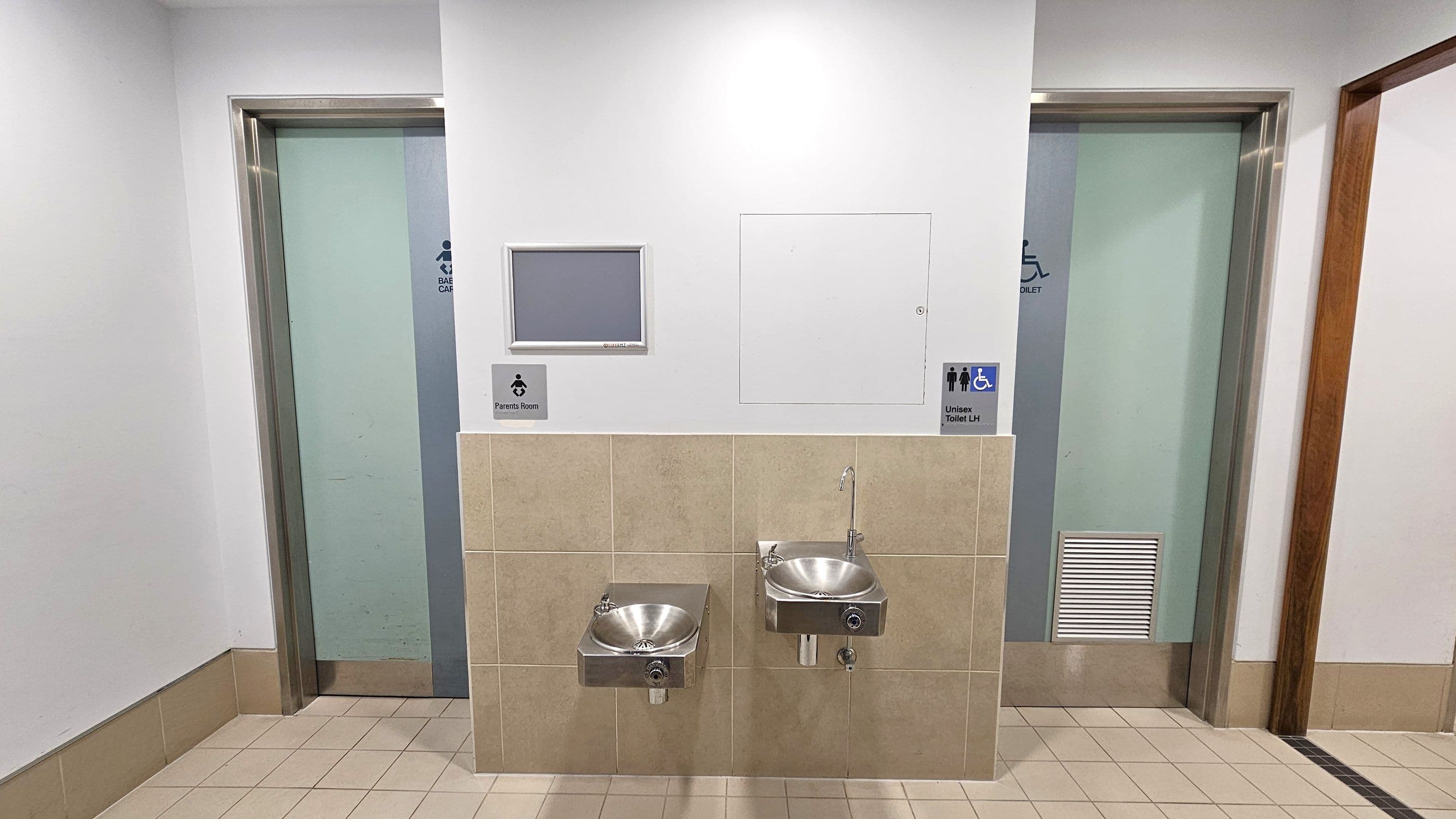 Two doors are visible, one on the left labelled "Baby Care" and another on the right labelled "Unisex Toilet LH" with accessibility symbols. Between them, there are two stainless steel drinking fountains mounted on a tiled wall. Above the fountains, there are signs for a "Parents Room" and the unisex toilet. The floor is tiled in a light colour, and the walls are painted white.