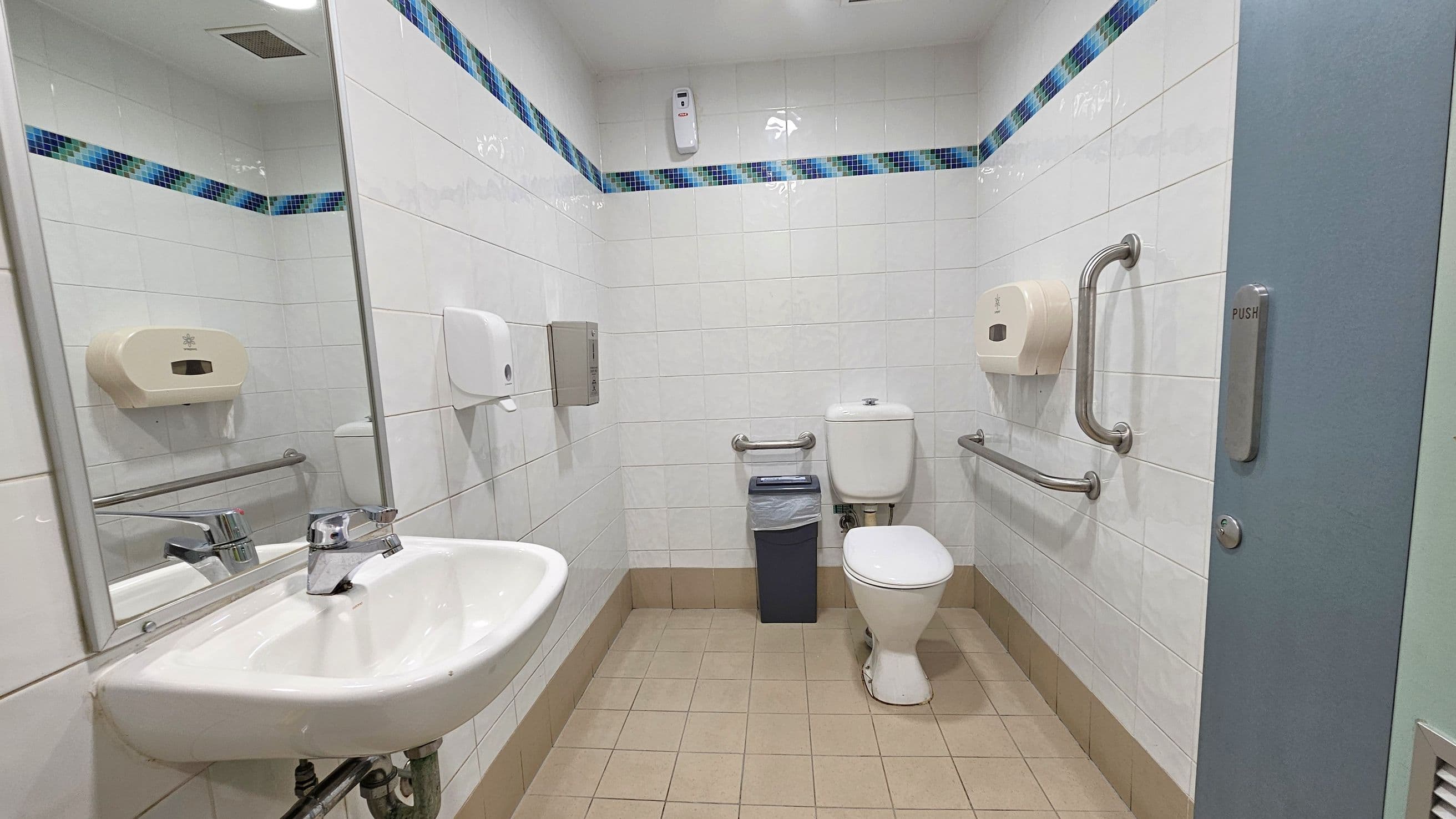 A public restroom with white tiled walls and a blue mosaic border. On the left, a sink with a mirror above it and a soap dispenser. In the centre, a toilet with grab bars on both sides and a small bin nearby. On the right, a door with a "PUSH" sign. The floor is tiled in a light brown colour.