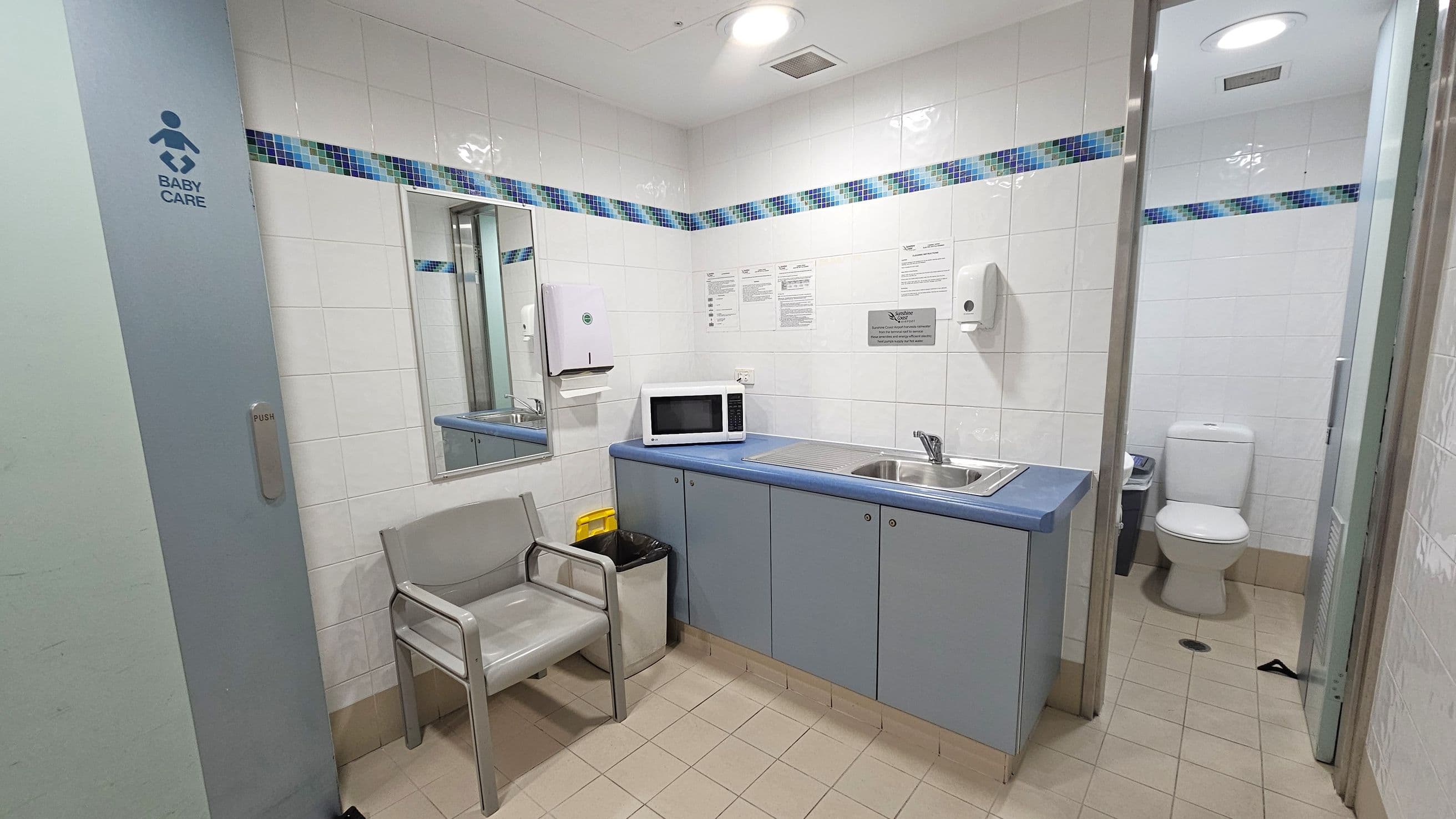 A baby care room with a tiled floor and white tiled walls featuring a blue and green mosaic border. On the left, a door marked "Baby Care" is partially open. In the centre, a grey chair is next to a bin and a yellow waste container. Above, a mirror is mounted on the wall. A countertop with a blue surface holds a microwave and a sink. On the right, a doorway leads to a toilet. The ceiling is white with recessed lighting.