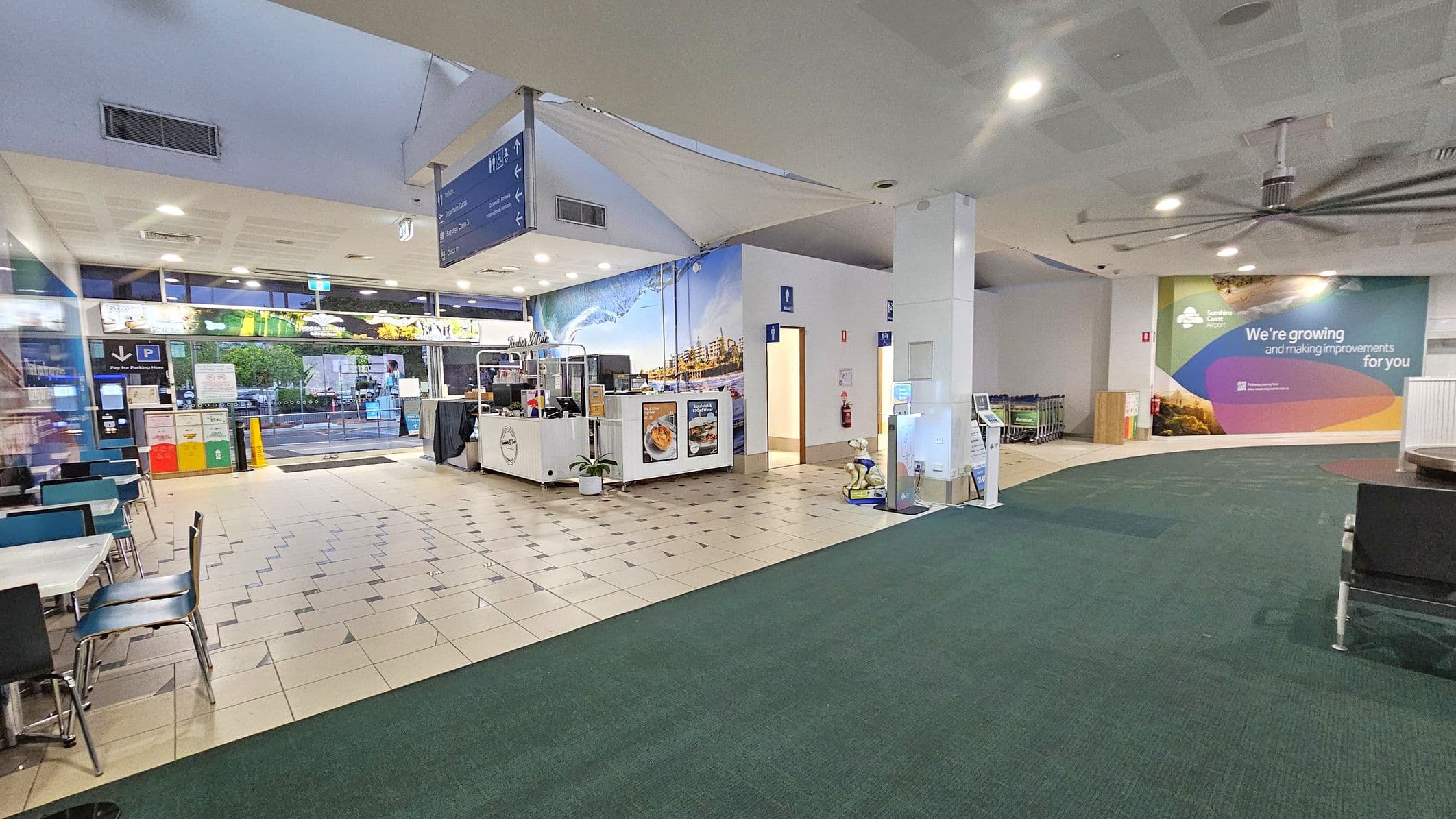 A spacious indoor area with a tiled floor transitioning to a green carpet. On the left, there are tables and chairs near a glass entrance with recycling bins and a parking sign. In the centre, a small café kiosk displays food images. To the right, a wall features a colourful mural with text about improvements. Nearby, there are restrooms and a row of luggage trolleys. The ceiling has recessed lighting and a large fan.