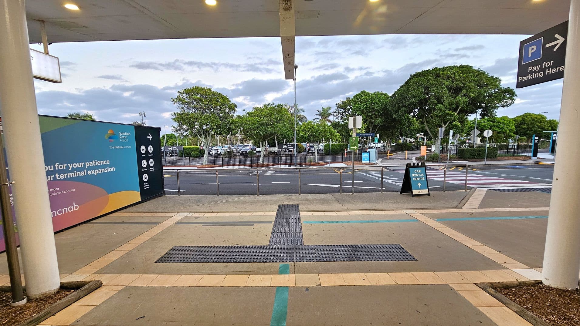 Under a covered area with a beige ceiling and spotlights, the foreground features a tiled floor with tactile paving. On the left, a large sign for Sunshine Coast Airport mentions terminal expansion. In the middle, a road with pedestrian crossings and a sign pointing to the car rental centre. The background has a parking area with several cars and lush green trees. On the right, a sign indicates parking payment.