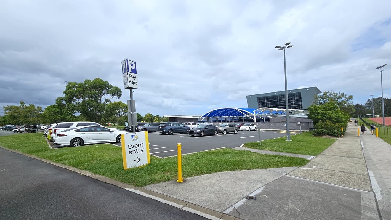 On the left, there is a car park with several parked cars and a sign that reads "Pay Here" with a large "P" symbol along with a sign indicating "Event entry" with an arrow pointing right. The right side features a pathway leading to a multi storey building with a blue dome shape shade structures. The background includes trees. The right shows a concrete pavement and a grassy area. Tall streetlights are also visible.