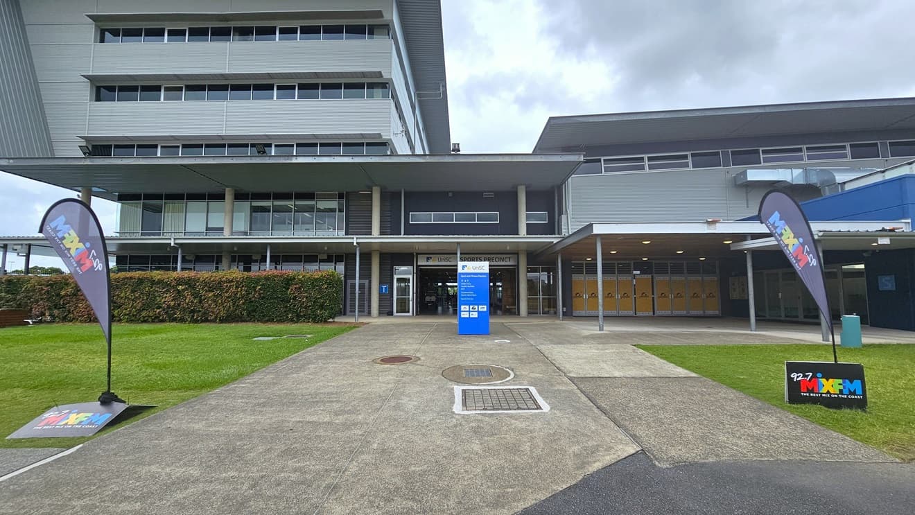 A multi-storey building is bordered at the base by a hedge. Centrally, a concrete pathway leads to an entrance marked by a blue sign and a metal grate on the ground. To the right, the building extends with large windows and a covered walkway. The upper floors, visible in the background, are clad in glass and metal panels. The foreground features concrete pavement and a grassy area, with feather banners and signage visible. Trimmed shrubs line the front to the left.