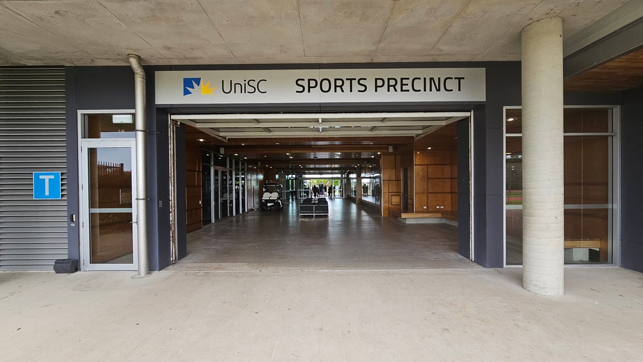 On the left, there is a grey wall with a blue "T" sign and a glass door. In the middle, an open entrance leads into a hallway with wooden panels and glass doors, with a sign above reading "UniSC Sports Precinct." The right side features a concrete pillar and a section of the building with dark walls. The background includes the interior of the sports precinct with visible lights and a small vehicle. The floor is concrete, and the ceiling is made of exposed concrete panels.