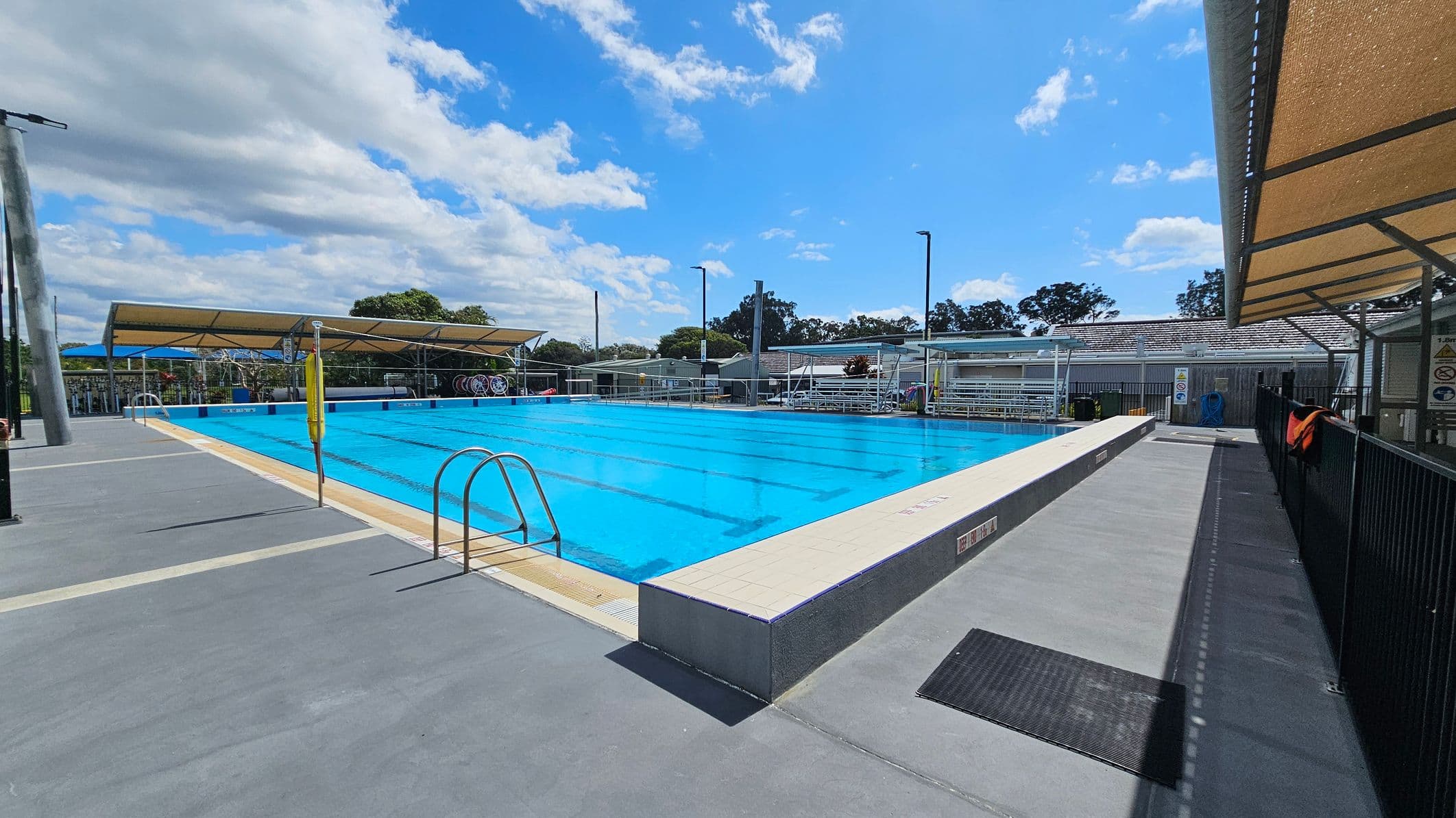 On the left, a row of large green shrubs adds a natural element beside a white building with vents and a smooth, light-coloured wall. To the right, a shaded area features a metal frame with a canopy, providing coverage over a walkway. The pool is visible in the middle, with clear blue water and a light-coloured edge. The ground is a clean, pale surface, and there are metal fences marking the pool area.