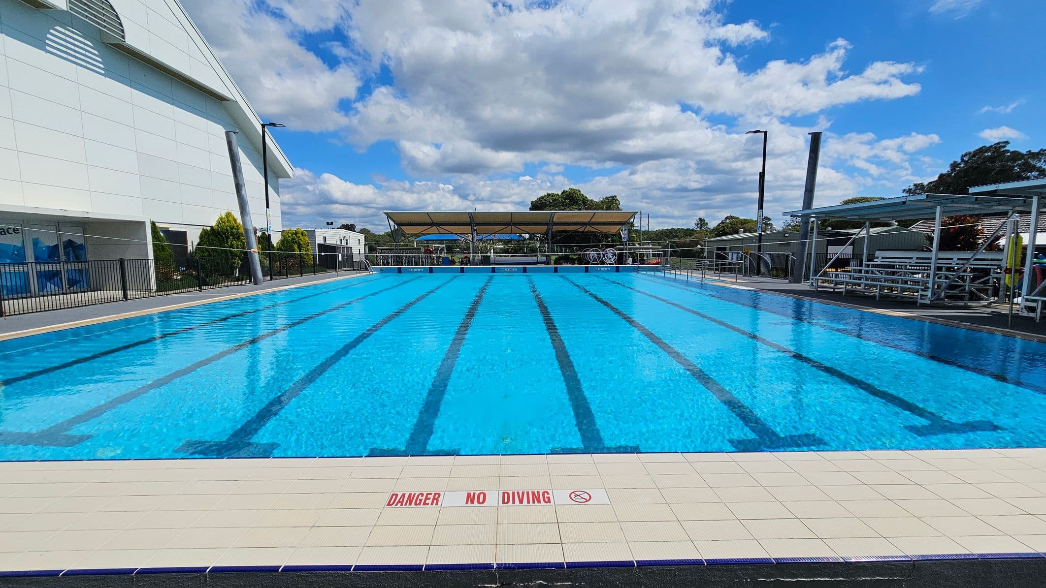 An outdoor swimming pool with clear blue water and marked lanes. The pool is surrounded by a tiled deck with a "Danger No Diving" warning. On the left, there's a large building with a slanted roof and some greenery. On the right, there are metal bleachers under a canopy. In the background, trees and a covered area with equipment are visible. The sky is partly cloudy.