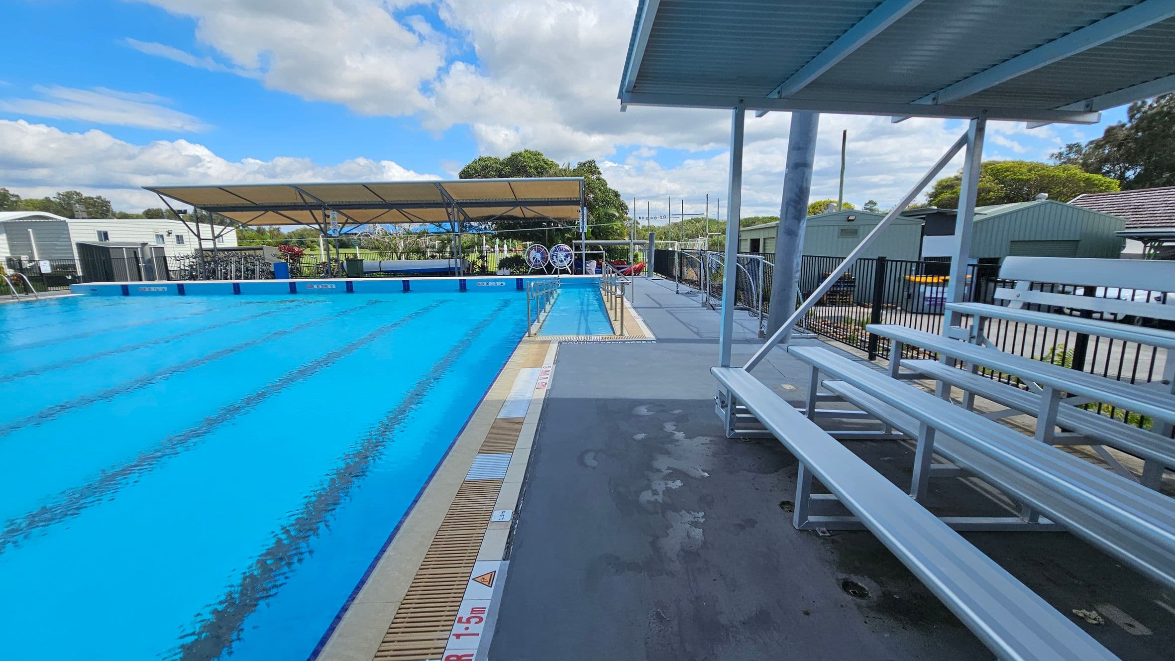On the left, there's a large swimming pool with clear blue water, bordered by a white building with a slanted roof. A yellow flotation device hangs on a pole by the poolside. On the right, there are metal bleachers under a corrugated roof, with a bin filled with colourful pool noodles. In the background, there are some plants and a fence, with a few structures visible beyond the pool area.