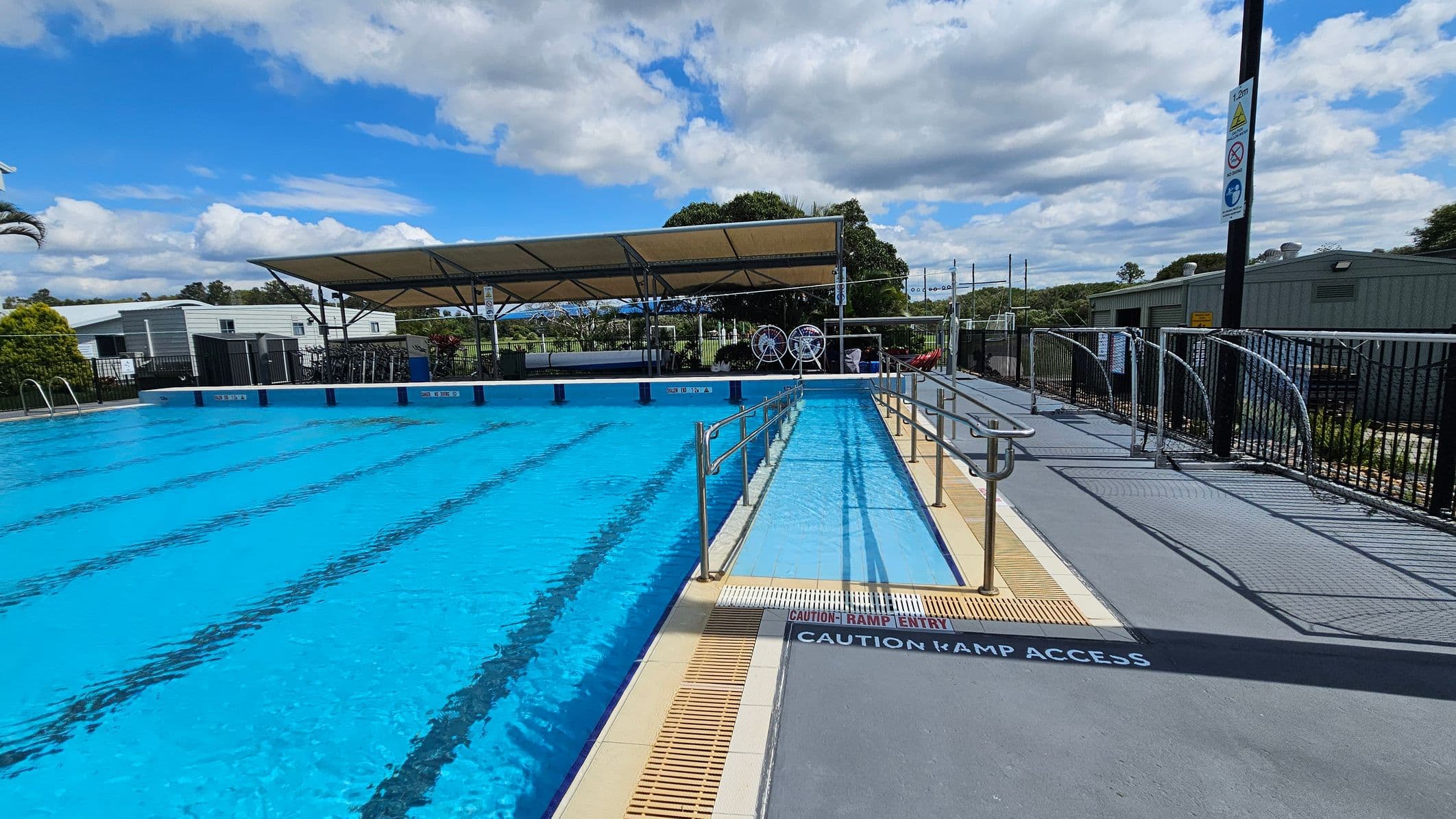 An outdoor swimming pool with clear blue water and lane markings. On the right, there's a ramp with metal railings leading into the pool, marked with "Caution Ramp Access." A shaded area with a canopy is in the background, along with some buildings and trees. The pool deck is grey with a black fence running along the perimeter. Signs are visible on a pole near the fence.
