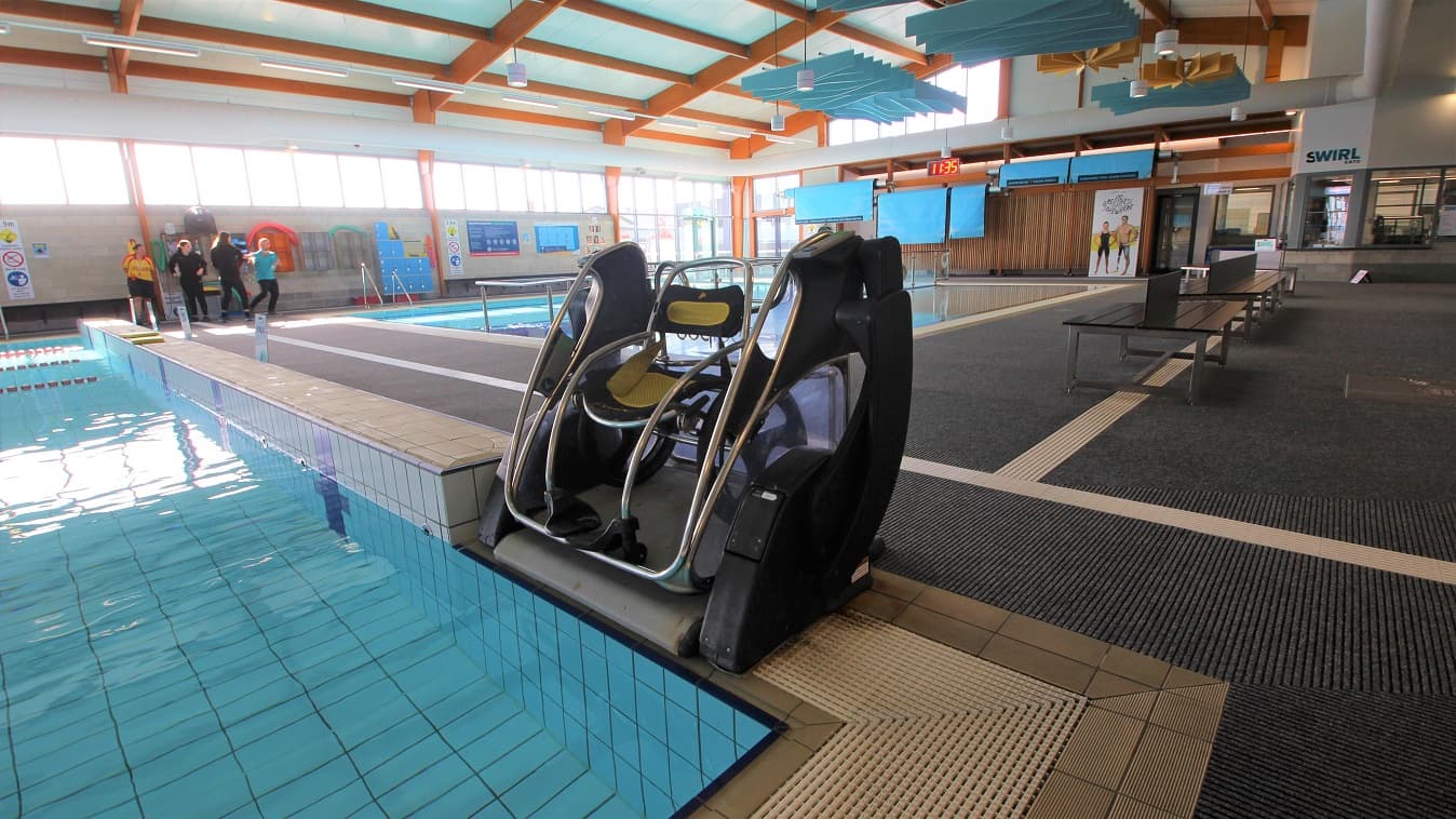 Aquatic wheelchair in the Aquatic facility at SWIRL. It is placed near the railed entrance ramp of a three-lane (6m x 12.5m) program pool that is 0.9m in depth. A digital clock can be seen placed on the upper wall of the facility. Separating the three-lane (6 m x 12.5m) program pool and the shallow pool found near the entrance door is a steel frame with a glass wall.
