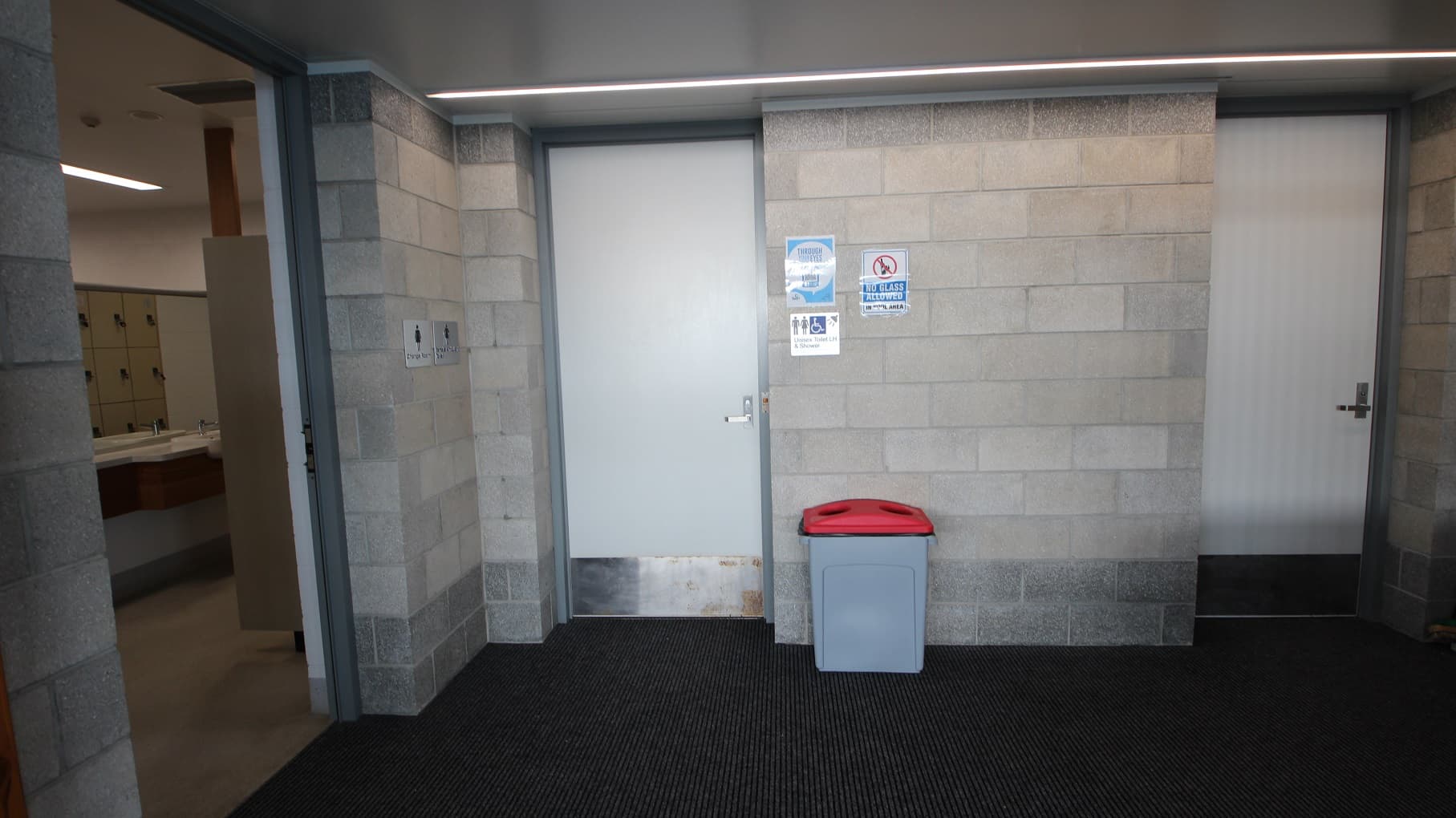 Unisex toilet and shower at SWIRL. A no glass allowed sign can be seen on the grey brick wall on the right side of the door. A grey rubbish bin with a red lid can be seen right outside the unisex toilet and shower room. Adjacent is an open door for toilets.
