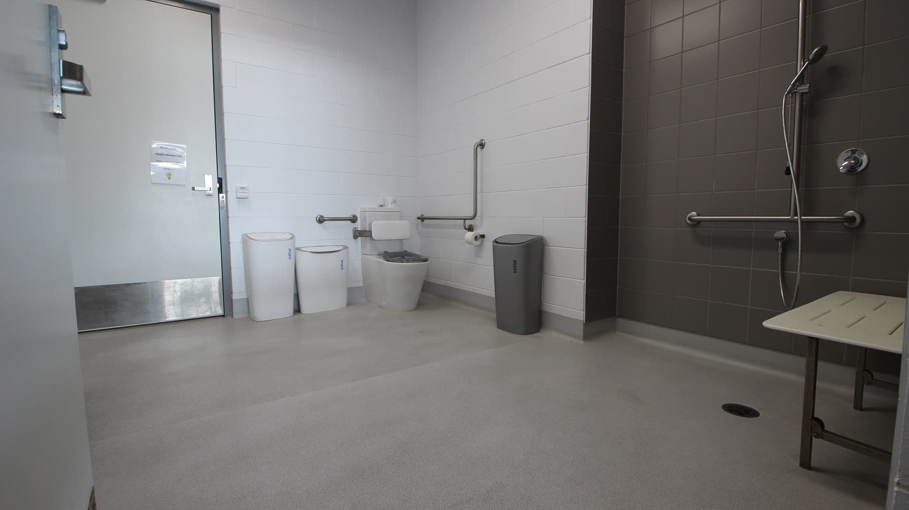 Inside look at the accessible change rooms at SWIRL. The shower area, equipped with grab bars and a handheld shower head, is painted grey. On the back is the toilet seat with a white cushion for the back. Mounted on the white tiled wall near it are grab bars and a toilet holder with a roll of toilet paper. On the focal point at the back is another door.