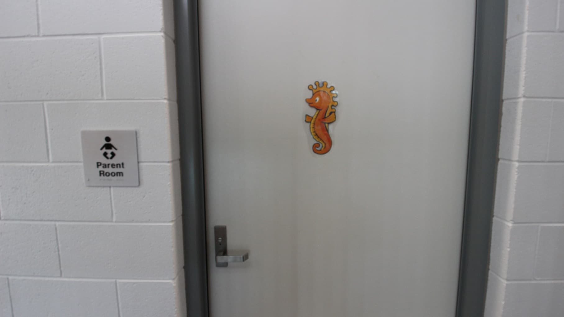 Parent room at SWIRL. It can be distinguished by the cute orange seahorse in the middle of the door and a white-painted brick wall. The door has a metal handle.