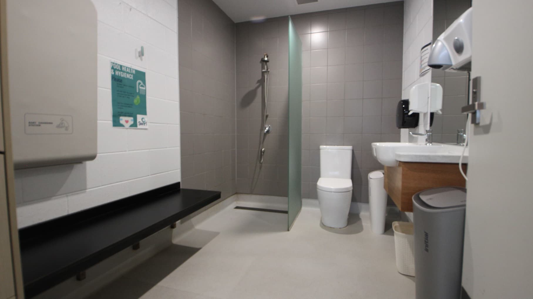 Inside the family change room at SWIRL. Mounted on the left white-painted brick wall is a black bench. The shower area and toilet seat are separated by a glass partition and the brick wall is painted in grey. On the right side are a wooden wash basin, a large mirror, tissue for hands, and a hand dryer