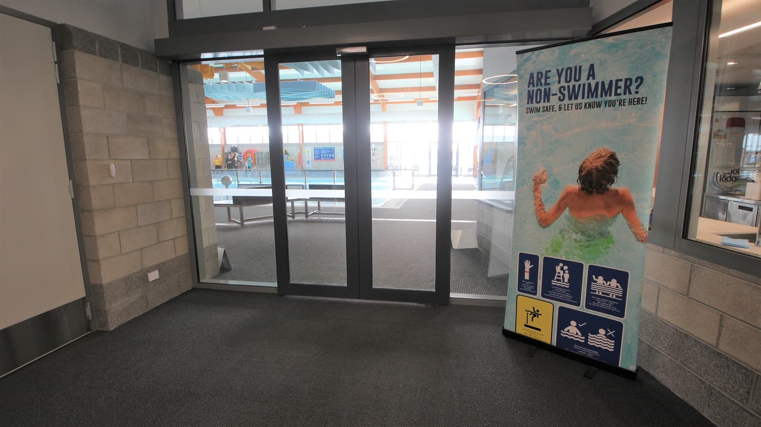 Automatic double glass entrance doors of the Aquatic facility inside SWIRL. The brick wall of this corridor has grey with a bit of dark portion at the bottom. On the right is a large poster with a picture of the child in a pool with the precautionary measures that need to be remembered by guests on the bottom.