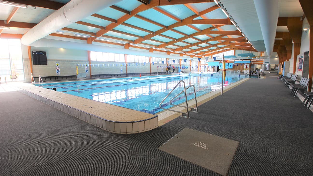 Opposite end of the 25-metre indoor pool that starts at 0.9m depth through to 1.8m depth with a 1.2m deep zone. The lanes are separated by red and blue floating ropes. There is a ladder found on both ends of the pool. This side of the pool has a string of hanging blue and white flags tied to opposite poles on the sides of the pool. Grey chairs can be seen on the right.