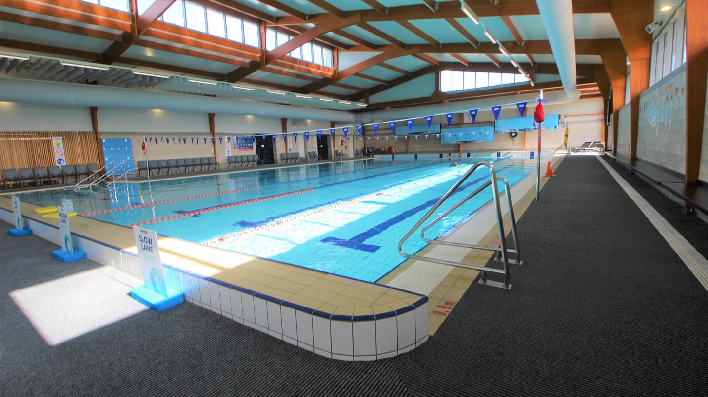 Opposite side of the stair entrance of the 25-metre indoor pool at SWIRL. There is a pool ladder on this side and white sign boards for the slow lane, medium lane, and fast lanes of the pool. Mounted on the white tiled wall on the right is a black seating platform.