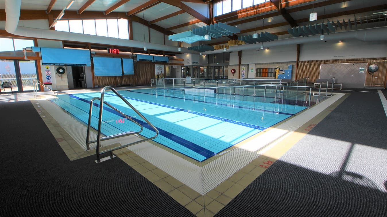 A closer look at the three-lane (6m x 12.5m) program pool that is 0.9m in depth that has a railed entrance ramp. There are two pool ladders on each end of the pool. Seen from this position is a digital clock placed on the upper wall of the facility. Separating the three-lane (6 m x 12.5m) program pool and the shallow pool found near the entrance door is a steel frame with a glass wall.