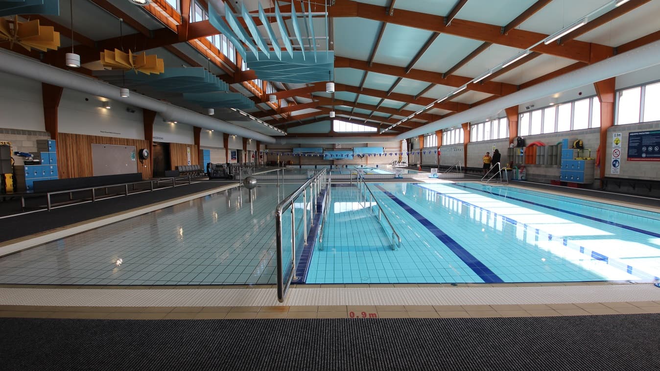 Central view of the three-lane (6 m x 12.5m) program pool and the shallow pool found near the entrance door is a steel frame with a glass wall. There are blue locker cabinets placed against the wall and grey-orange storage cabinets for bags mounted on the right of the three-lane pool.