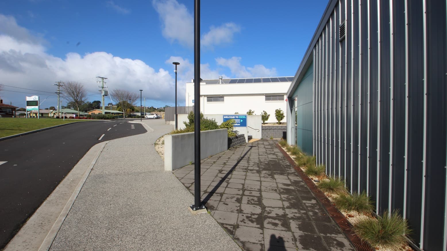 Crossing the zebra lanes and turning right off the smooth cemented pathway with a black lamp post is a grey tile floor to the SWIRL entrance. Tall grasses are linearly arranged against the light green and black segmented walls of the facility. Heading straight from this pathway and to the left is a Welcome to SWIRL sign placed on the short white wall next to a staircase with metal handrails.