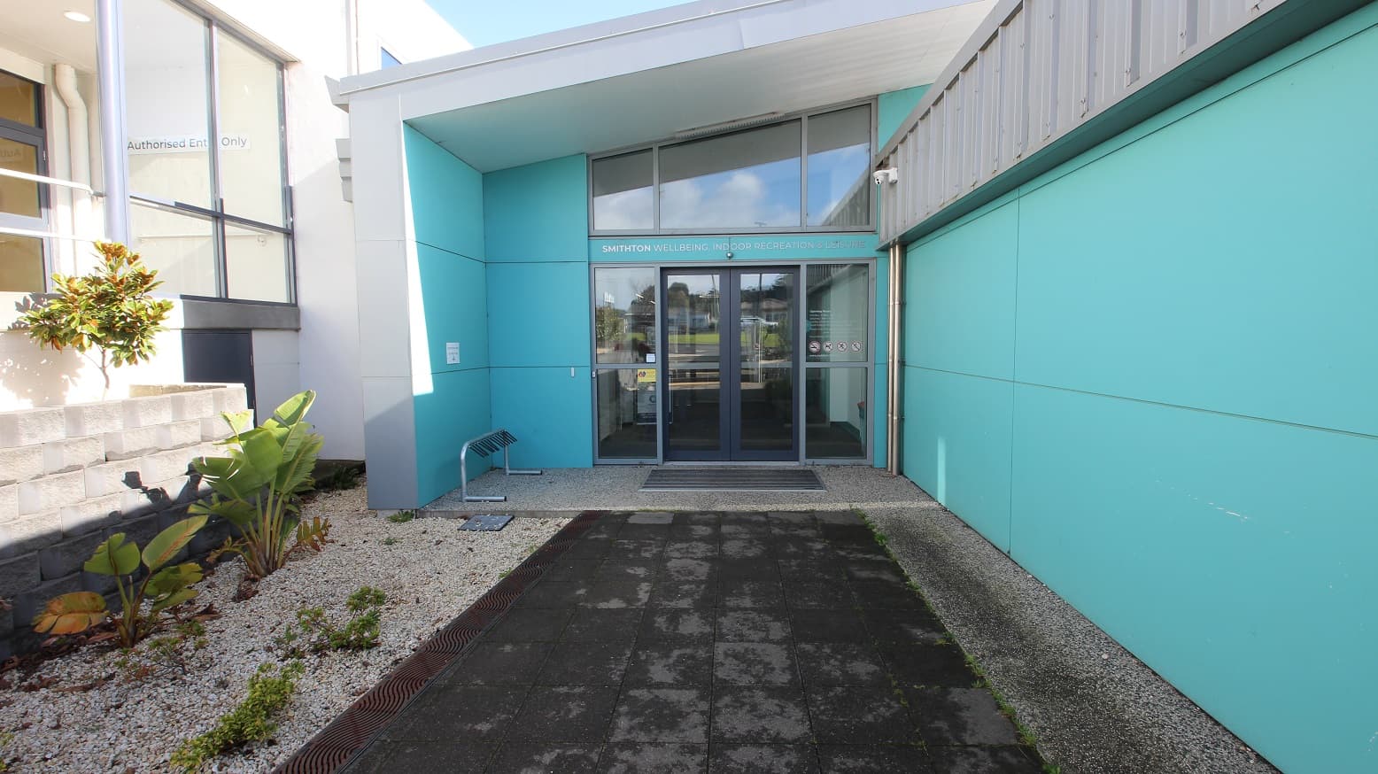 Grey tiled pathway leading to the entrance automatic, double glass doors of SWIRL. There is a type of plant from the banana family due to its leaves in the garden box. Prohibited things inside the facility are displayed on the right glass door (no smoking sign, no pets allowed, no running, etc). On the top of the glass doors is SMITHTON WELLBEING INDOOR RECREATION & LEISURE. Found on the wall at the end of the entrance ramp of the white building is an Authorised Entry Only sign.