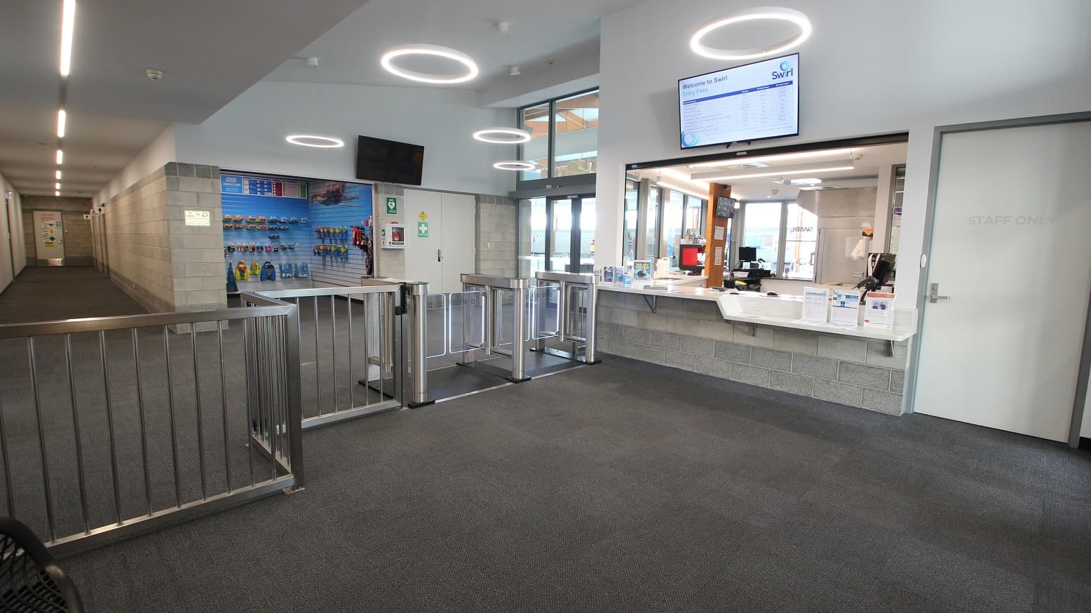 Turnstiles, including an accessible turnstile that leads to a small retail space selling a range of aquatic gear. Staff can go to the reception area through a white door with a STAFF  ONLY sign. On the upper white wall of the reception desk is a television showing information about SWIRL. The retail space has a grey brick wall.  There is a corridor found on the left, next to the retail space.